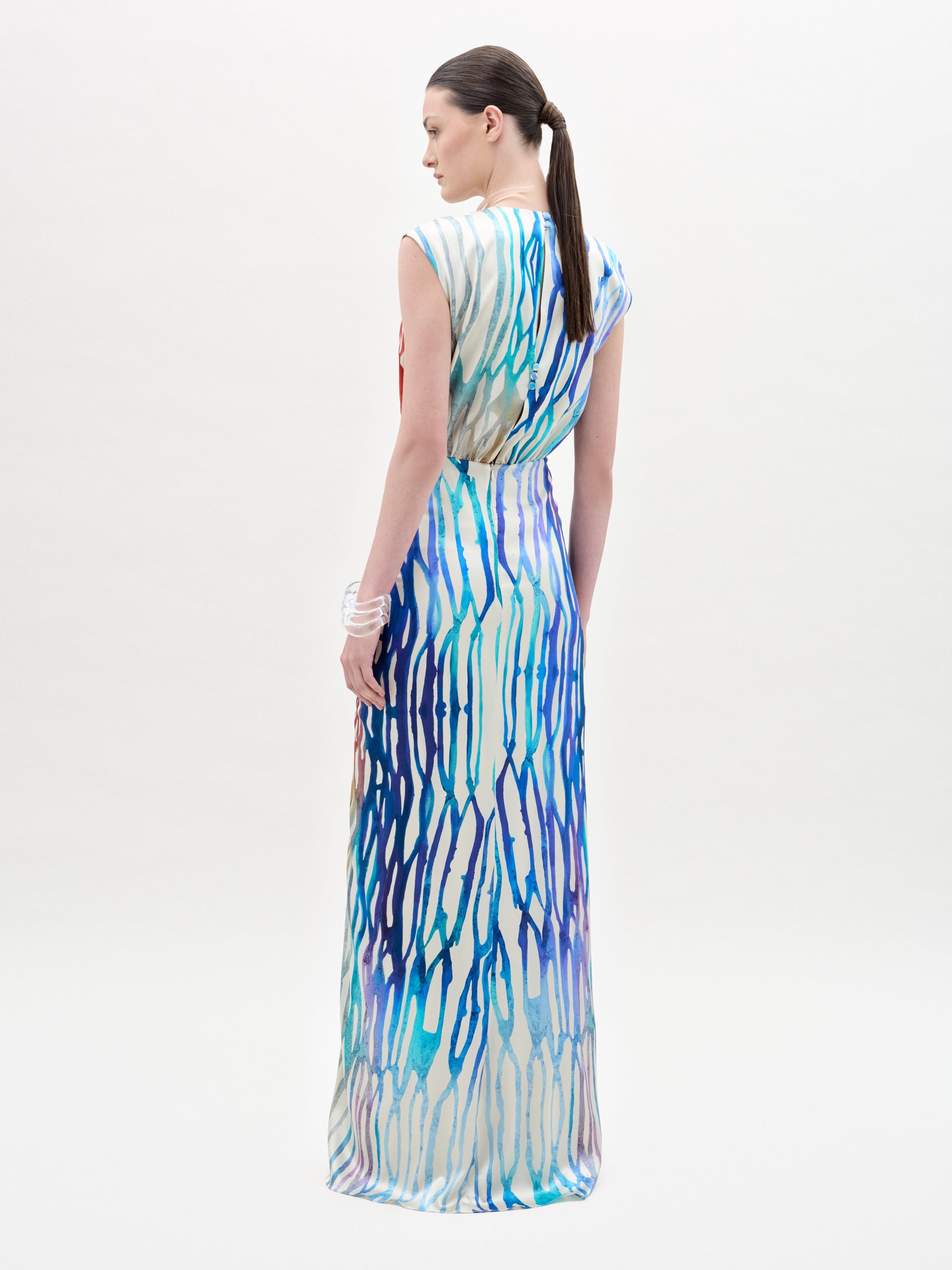 A woman with a low ponytail models the Samaira Dress by SVT DESIGNS, a sleeveless maxi with a ruched waist and blue, purple, and white abstract pattern. She wears a white bracelet on her left wrist against a plain white background.