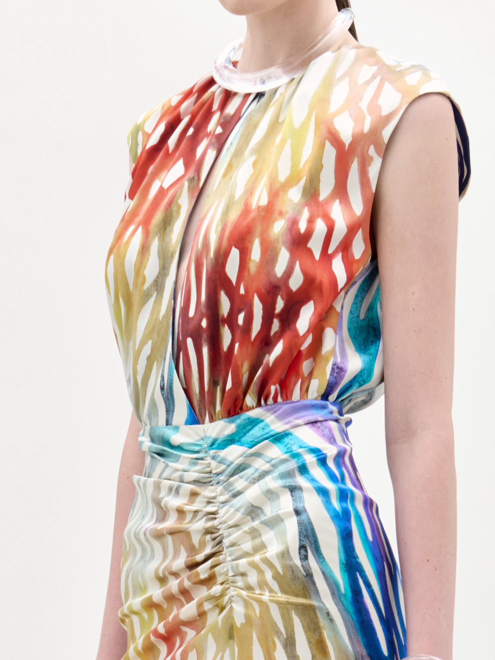 A woman models the Samaira Dress by SVT DESIGNS, a sleeveless, high-neck dress with a ruched waist. Made from Italian fabric, it features an abstract pattern in red, yellow, orange, blue, and white. Shown from shoulders to mid-thigh.