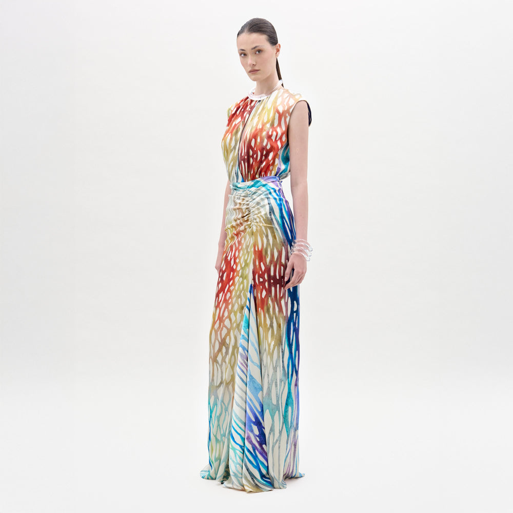 A woman poses against a white background in the Samaira Dress by SVT DESIGNS—a sleeveless, floor-length maxi with a vibrant red, blue, yellow, and white abstract print. Her dark hair is pulled back and she wears bracelets on her left wrist.