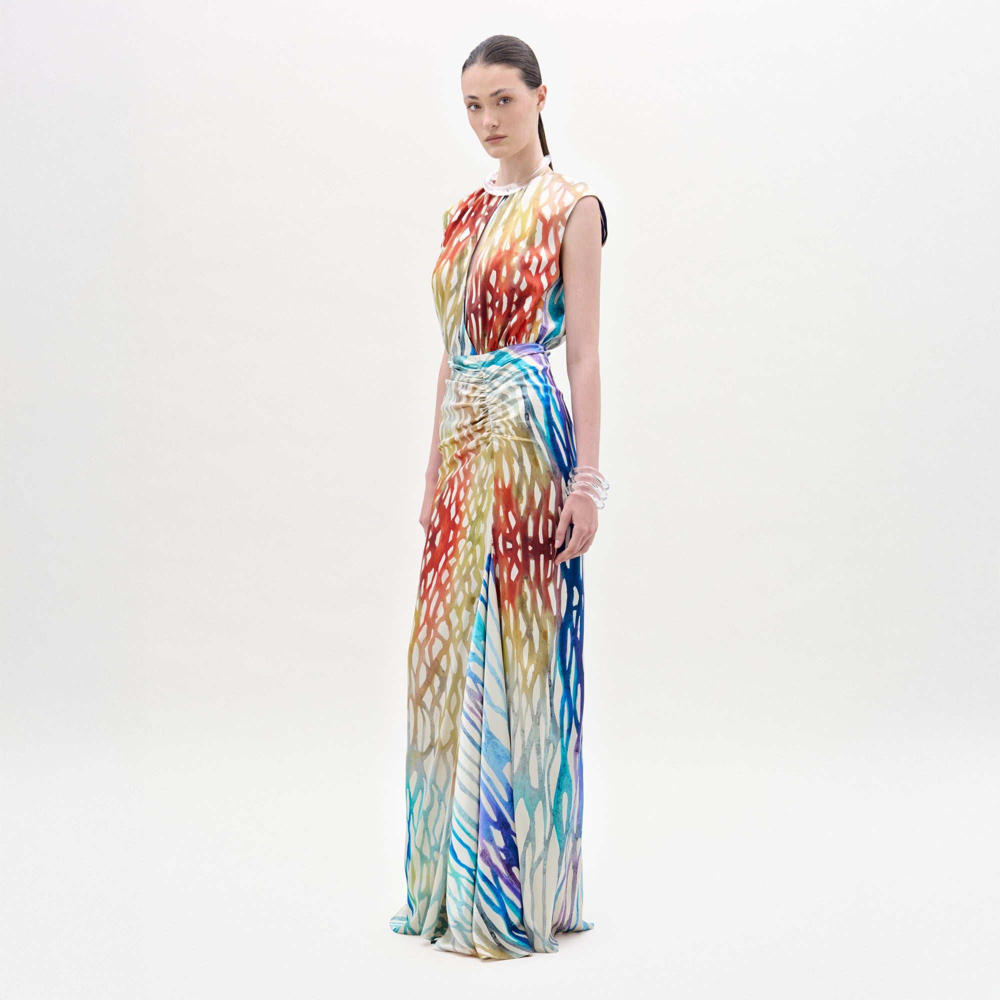A woman poses against a white background in the Samaira Dress by SVT DESIGNS—a sleeveless, floor-length maxi with a vibrant red, blue, yellow, and white abstract print. Her dark hair is pulled back and she wears bracelets on her left wrist.