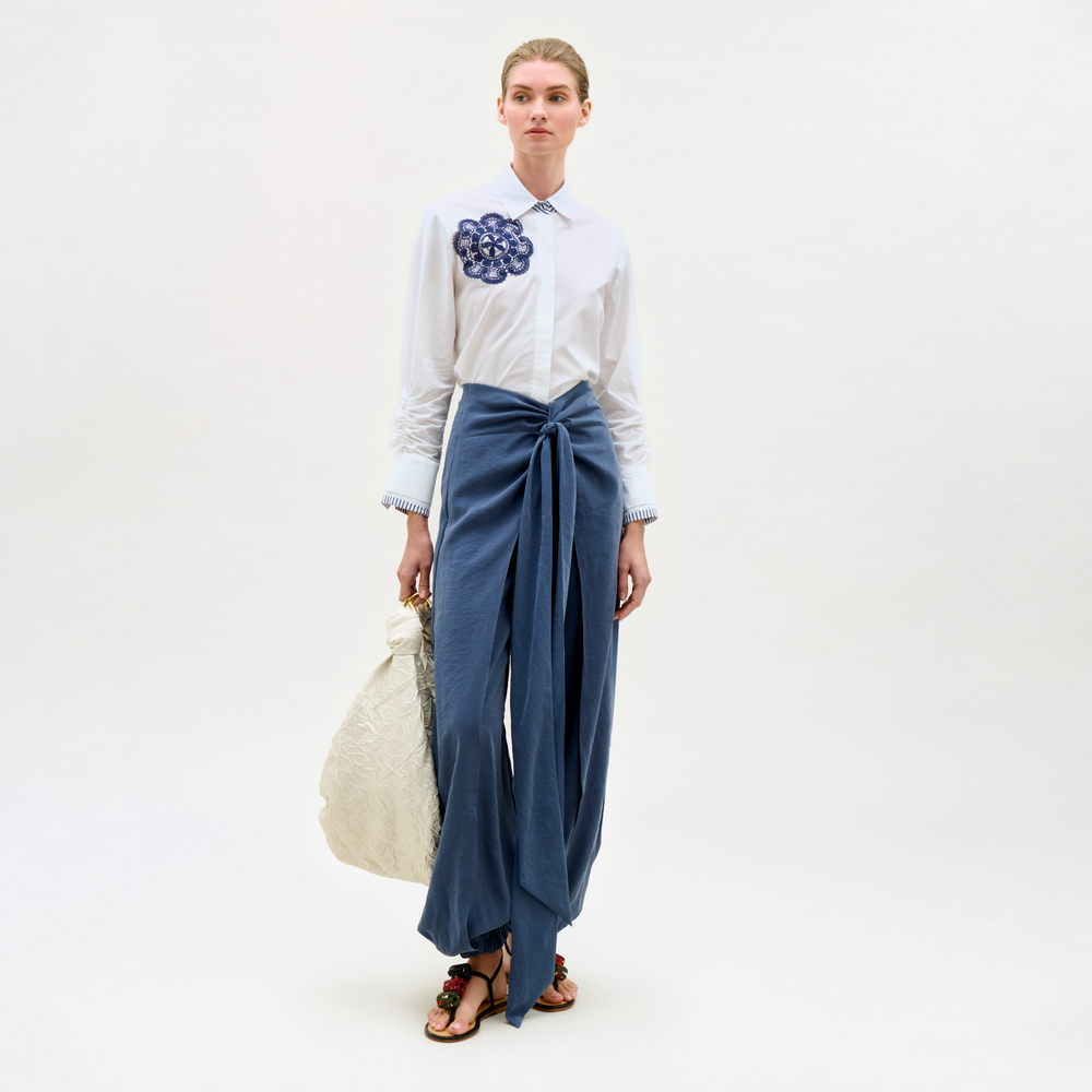 Woman wearing a white shirt with a floral design and blue pants, holding a beige bag on a white background