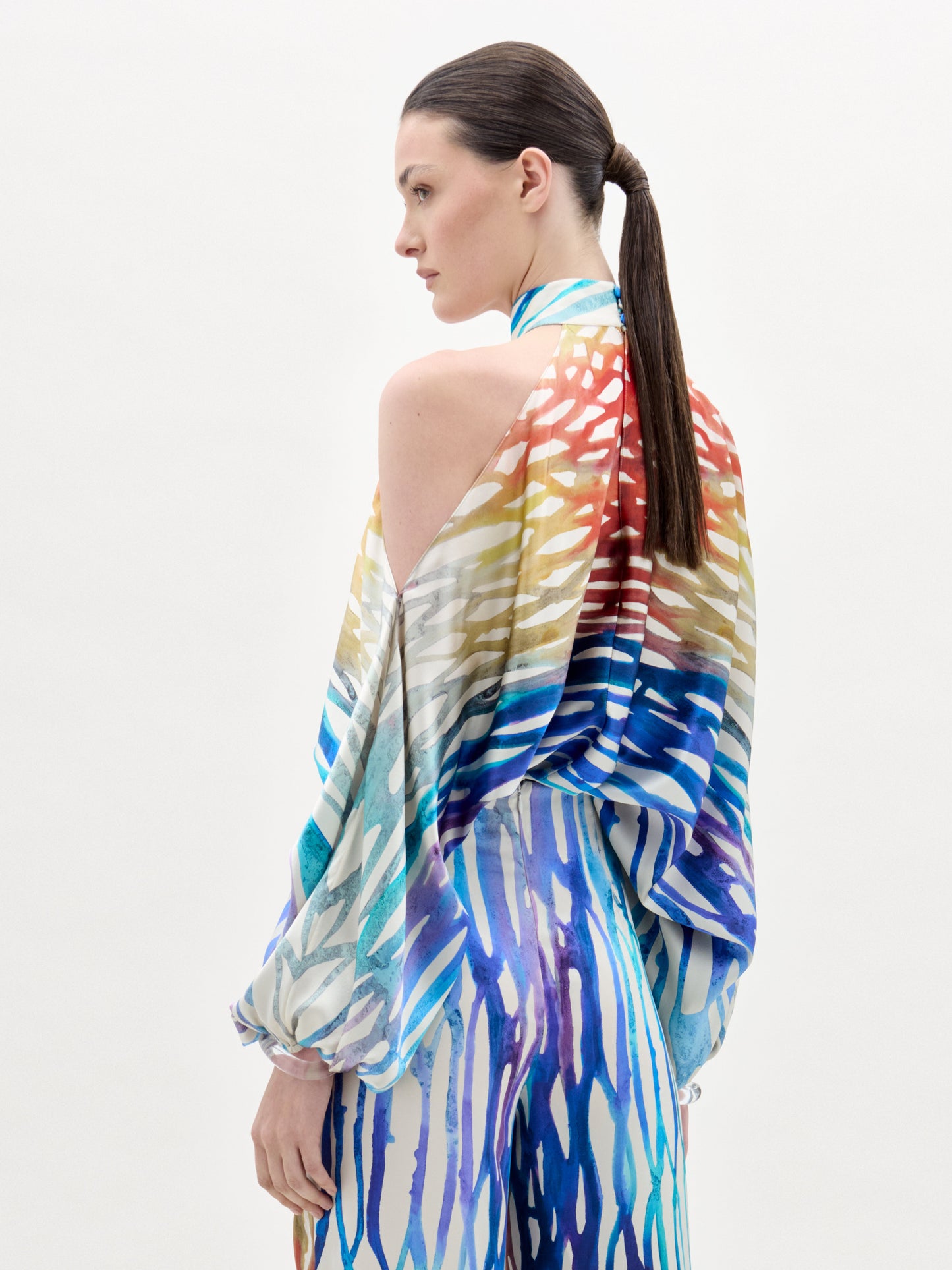A person with long, straight brown hair in a low ponytail wears the Safira Blouse by SVT DESIGNS—an abstract print top with open shoulders and loose sleeves featuring red, yellow, blue, and white stripes. The background is plain white.