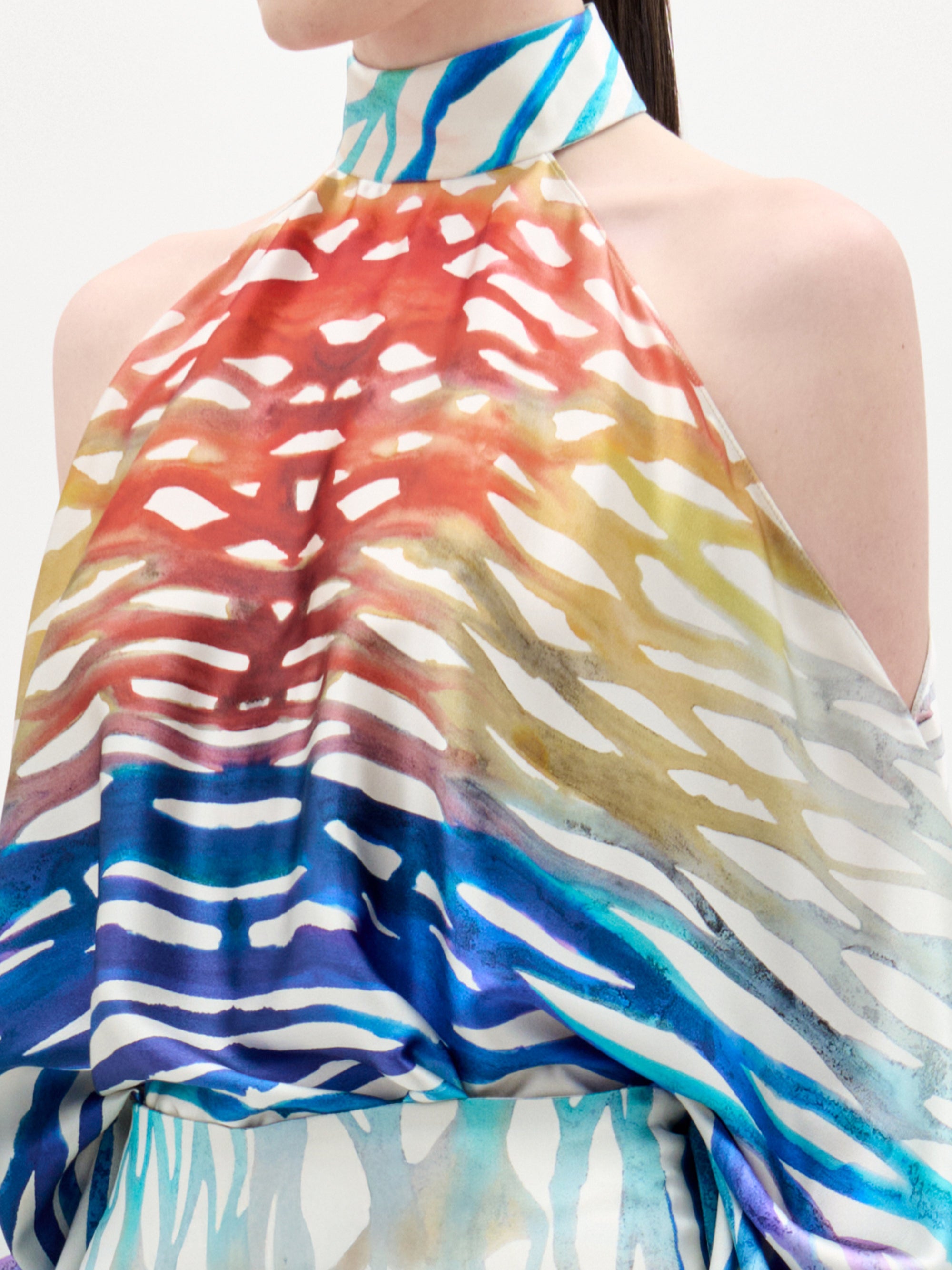 A person wearing the Safira Blouse by SVT DESIGNS, a sleeveless, high-neck top with a colorful abstract print in red, yellow, blue, and white. Only the torso and part of the face are visible.