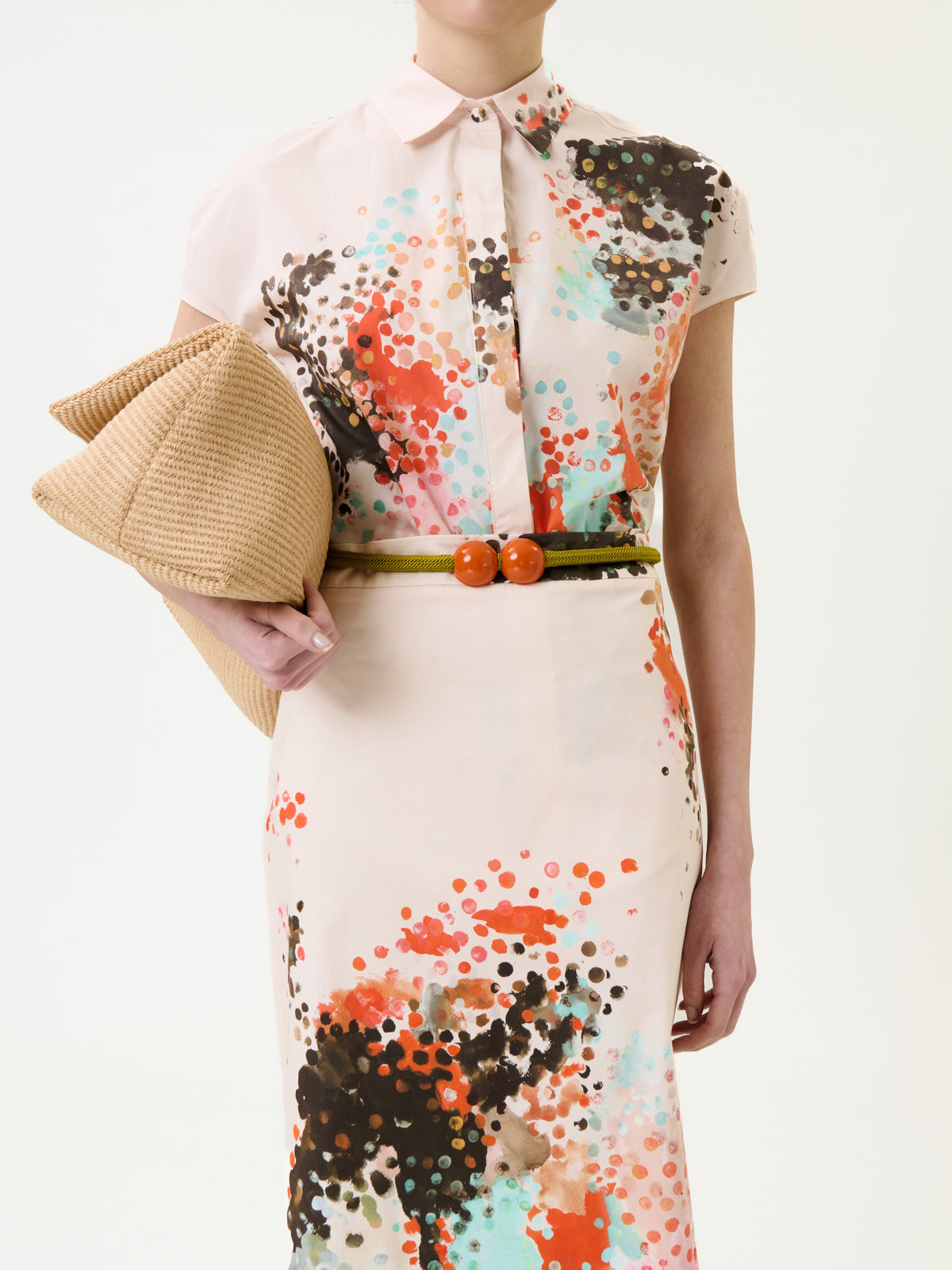 Woman in the Maribel Shirt, a short-sleeved, button-up dress with colorful abstract dots and a thin belt featuring two orange spheres at the center, holding a beige textured clutch. Her face is not visible.