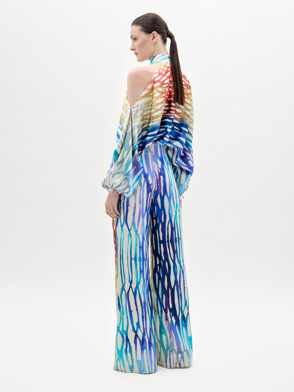 A woman with a sleek ponytail stands facing away in SVT DESIGNS' Andie Pant—high-waisted, wide-leg pants featuring a colorful wavy abstract pattern in red, blue, yellow, and purple. The background is plain white.