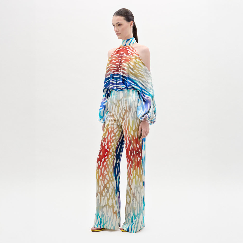 A woman stands against a white background wearing the Andie Pant by SVT DESIGNS, featuring loose, wide-leg pants made from Italian fabric with bold red, blue, yellow, and white abstract prints.