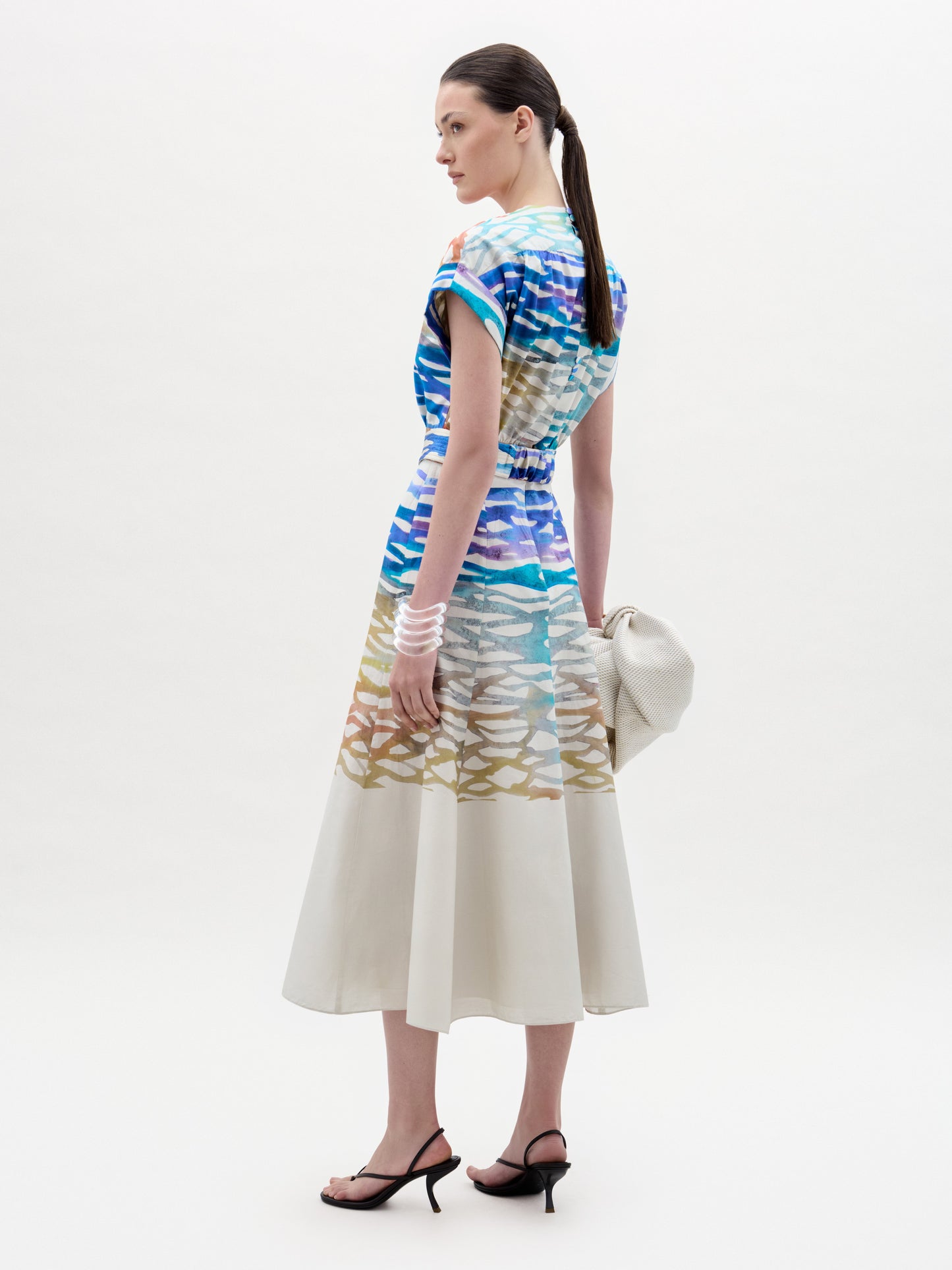 A woman stands slightly turned, wearing the SVT DESIGNS Sakura Dress—a colorful patterned midi with an A-line skirt—paired with strappy black sandals, a white bracelet, and a textured white clutch against a plain light background.