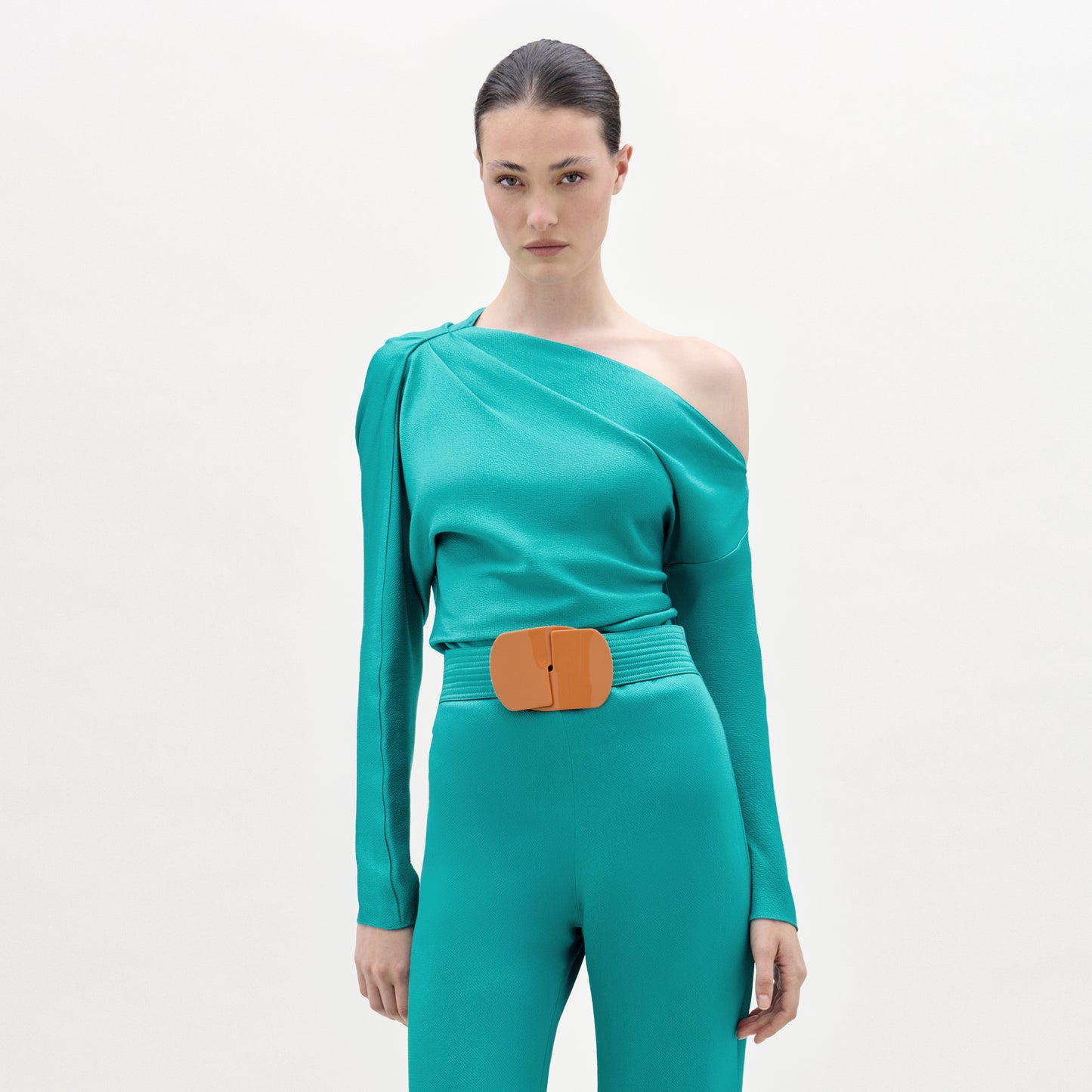 Model wearing a teal one-shoulder top and pants with a wooden belt on a light gray background