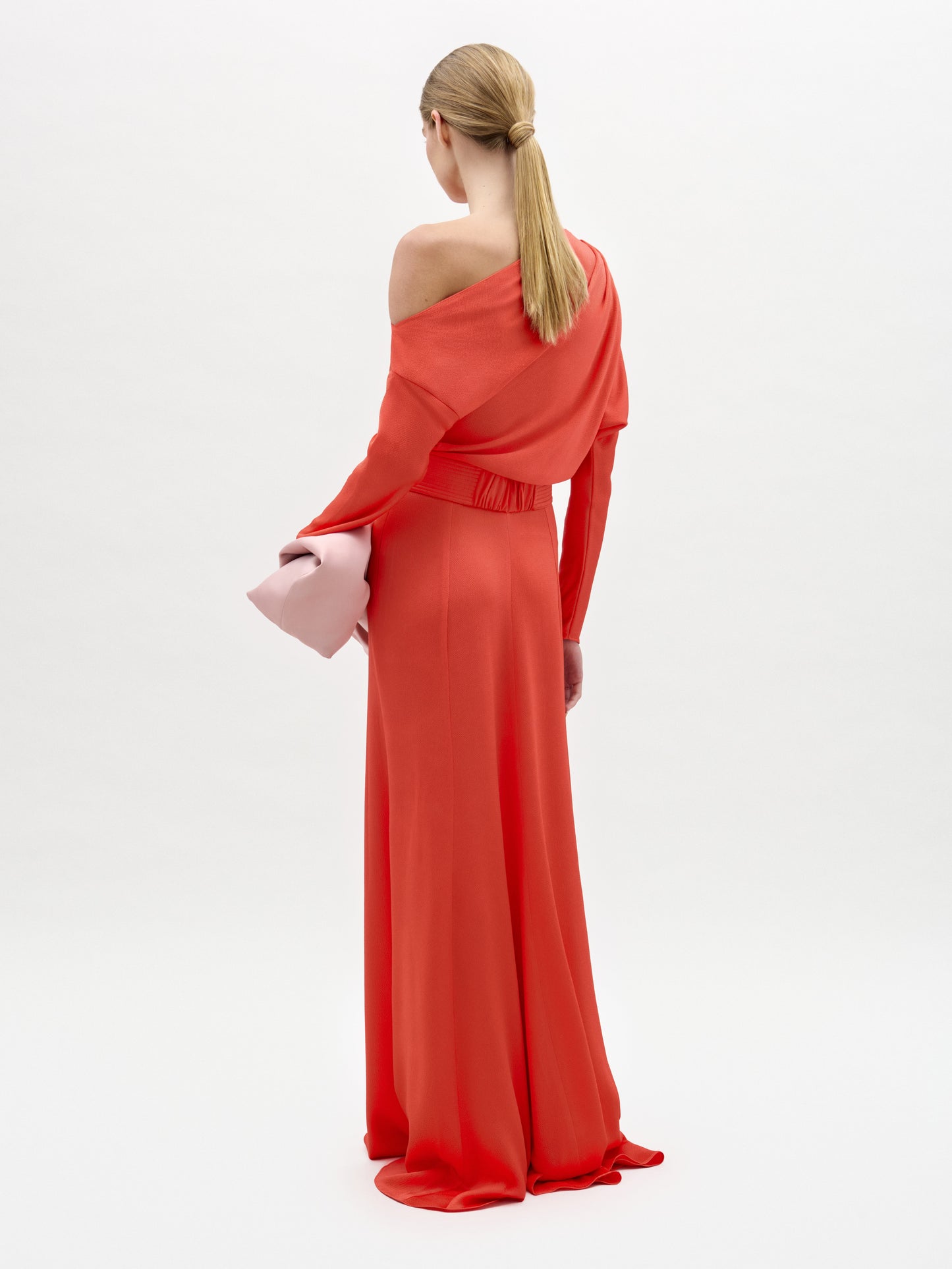 A woman with blonde hair in a low ponytail stands against a white background, facing away from the camera. She wears the SVT DESIGNS Noelia Dress, a long, bright red one-shoulder maxi, and holds a light pink clutch.