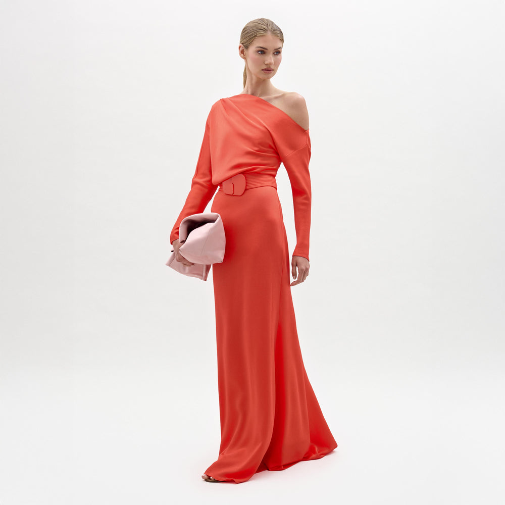 A woman poses against a plain white background wearing the SVT DESIGNS Noelia Dress—a bright coral maxi dress with an elegant one-shoulder neckline and belted waist—while holding a large pale pink clutch.