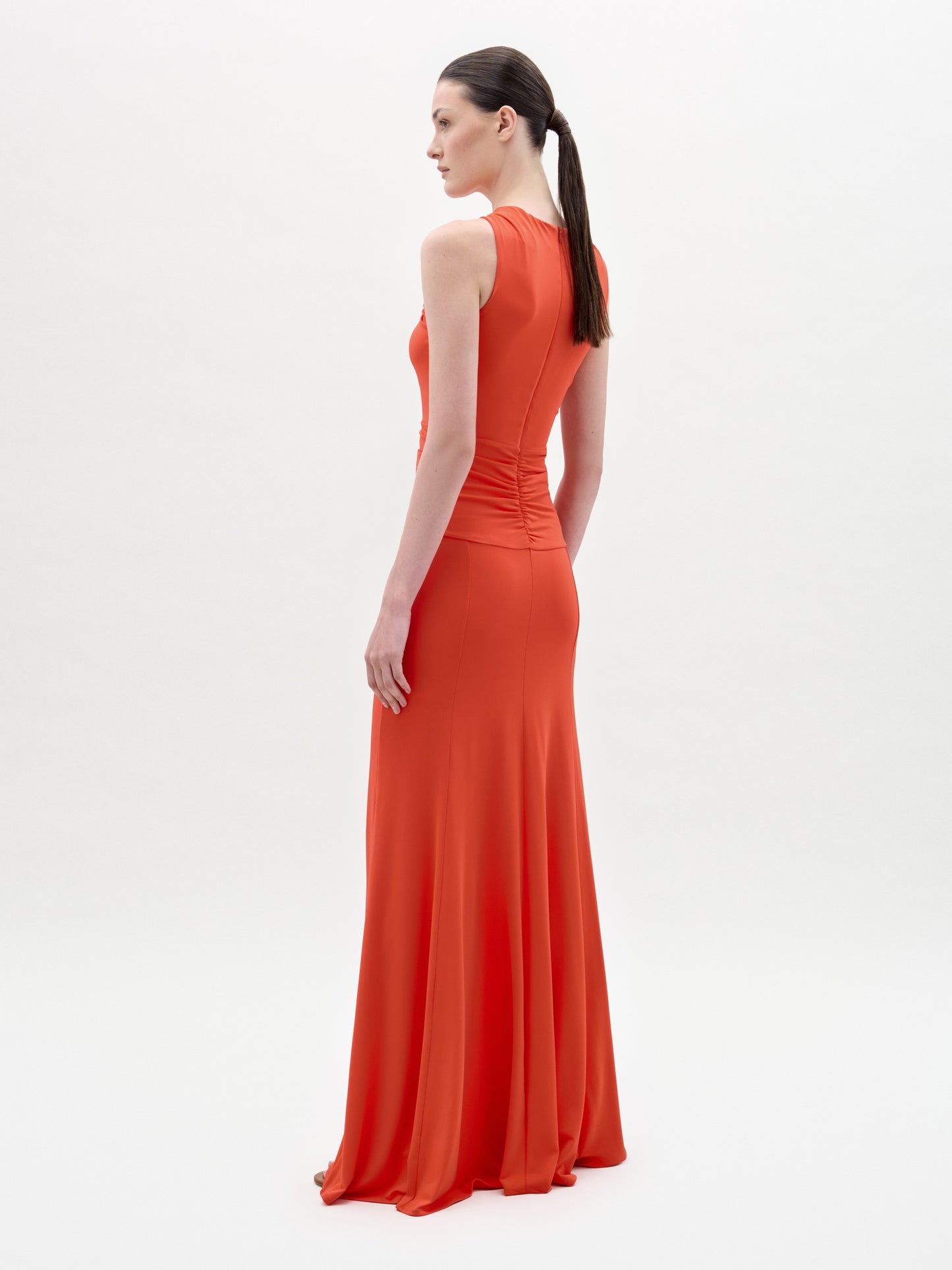 A woman with long dark hair in a ponytail stands in profile, wearing the sleeveless orange Naga Dress by SVT DESIGNS. The dress features a column silhouette. The background is plain white.