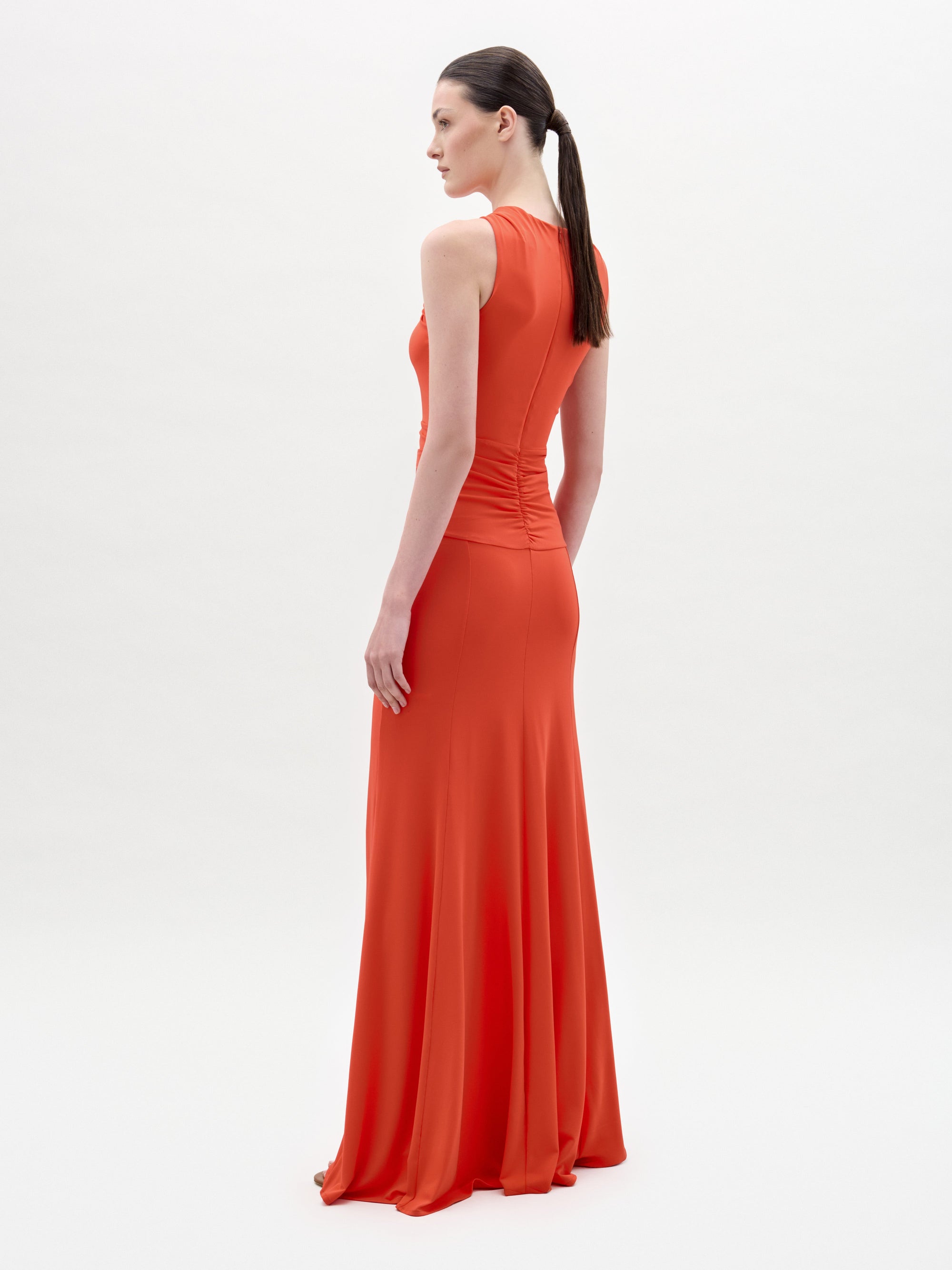 A woman with long dark hair in a ponytail stands in profile, wearing the sleeveless orange Naga Dress by SVT DESIGNS. The dress features a column silhouette. The background is plain white.