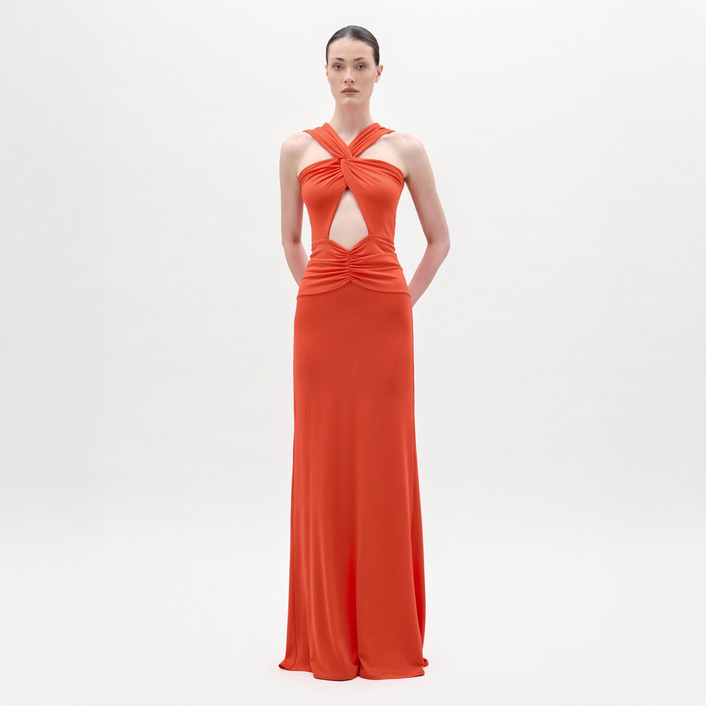 A person poses against a white background wearing the SVT DESIGNS Naga Dress—a sleeveless, floor-length bright orange maxi with a crisscross halter neckline, front cutouts, and a sleek column silhouette.