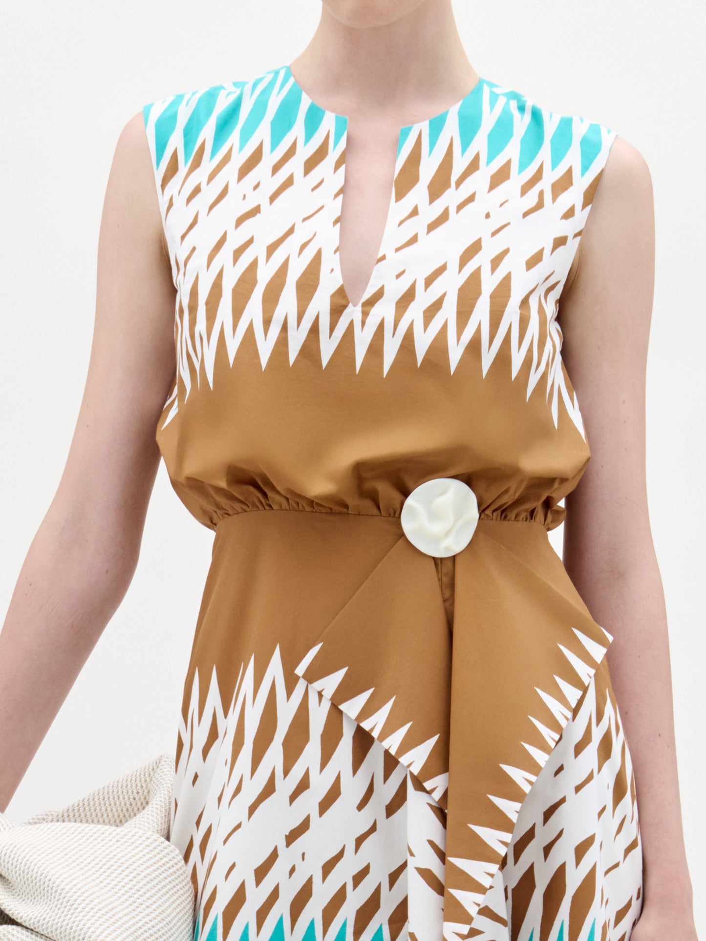A person wearing the Kendall Dress by SVT DESIGNS—a sleeveless, fit-and-flare midi dress with a geometric brown, white, and turquoise pattern, a notched neckline, oversized waist button, and asymmetrical ruffle detail.