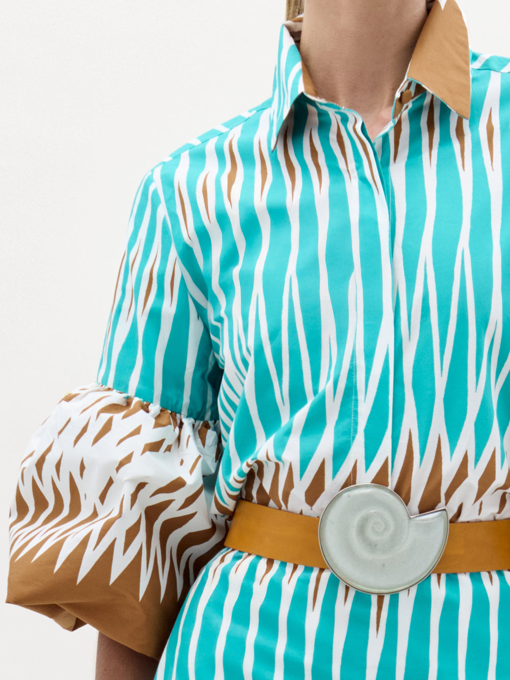 A person wears the Helina Blouse by SVT DESIGNS, featuring blue, white, and brown zigzag patterns, 100% cotton bell sleeves, and a mustard yellow belt with a large white shell-shaped buckle. Only the upper torso is visible.