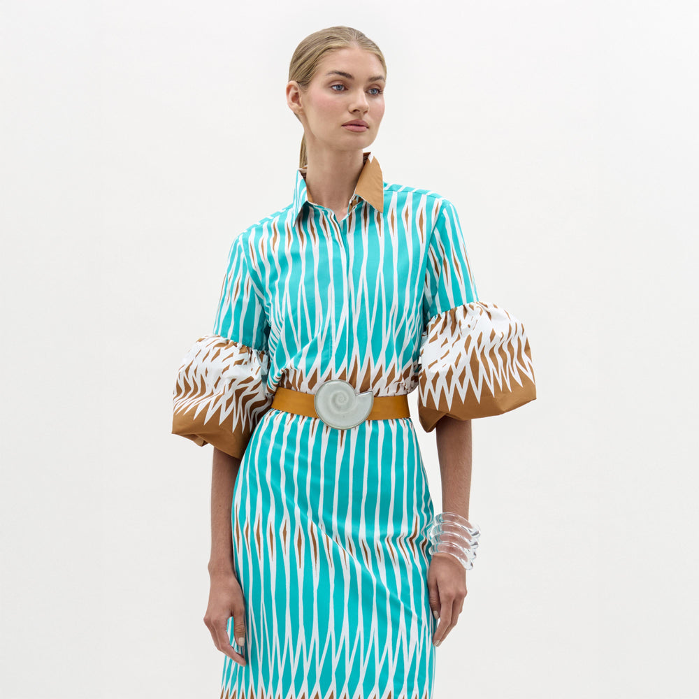 A woman stands against a plain background wearing the Helina Blouse by SVT DESIGNS, featuring turquoise and white stripes, 100% cotton fabric, wide puff sleeves with patterns, a tan collar, and styled with clear bangles.