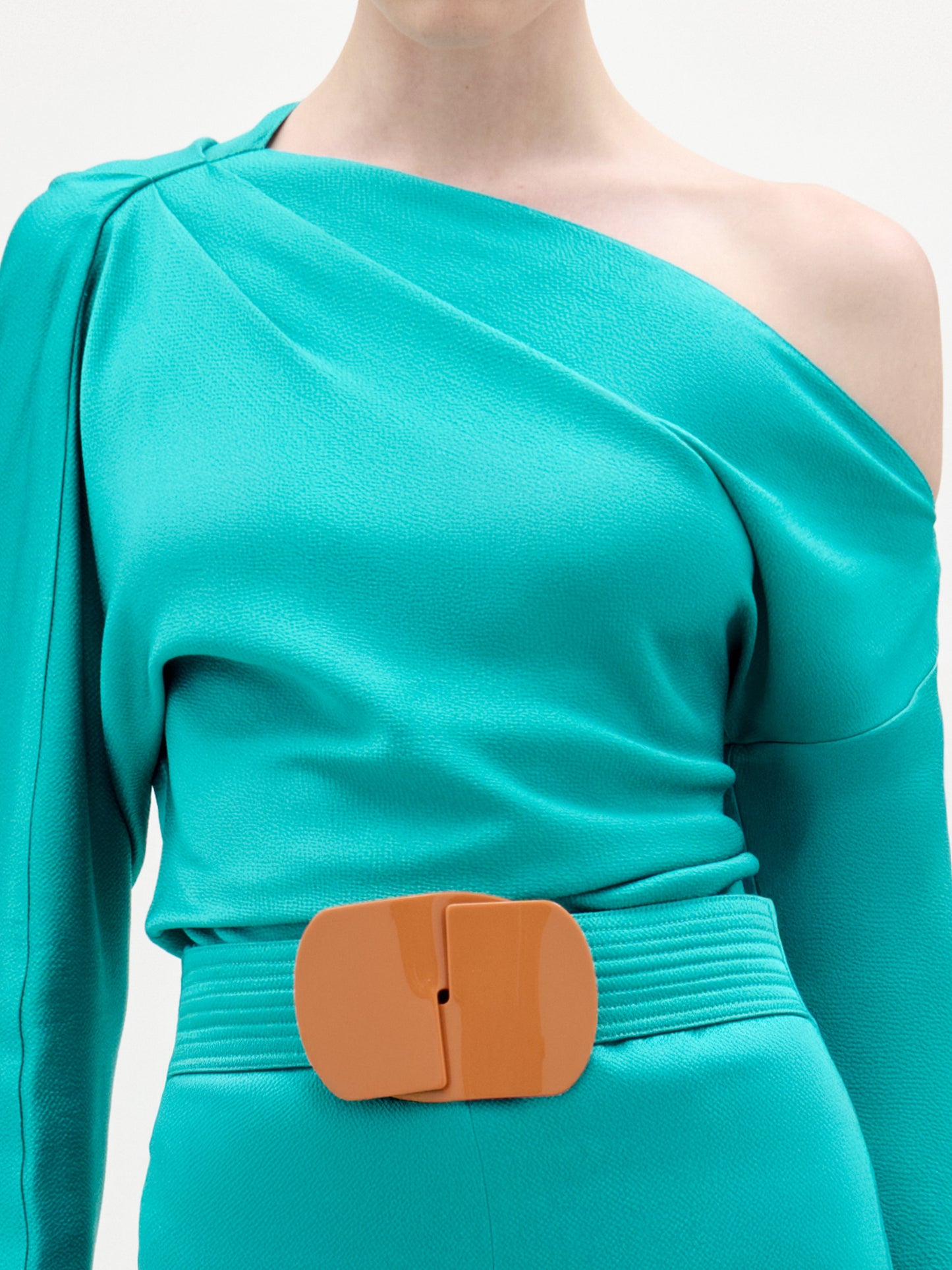 A person wears the SVT DESIGNS Viola Blouse in teal, featuring an asymmetrical one-shoulder neckline, paired with matching high-waisted pants and an orange rectangular belt. The sleek Italian fabric stands out. The person's head is cropped from the image.