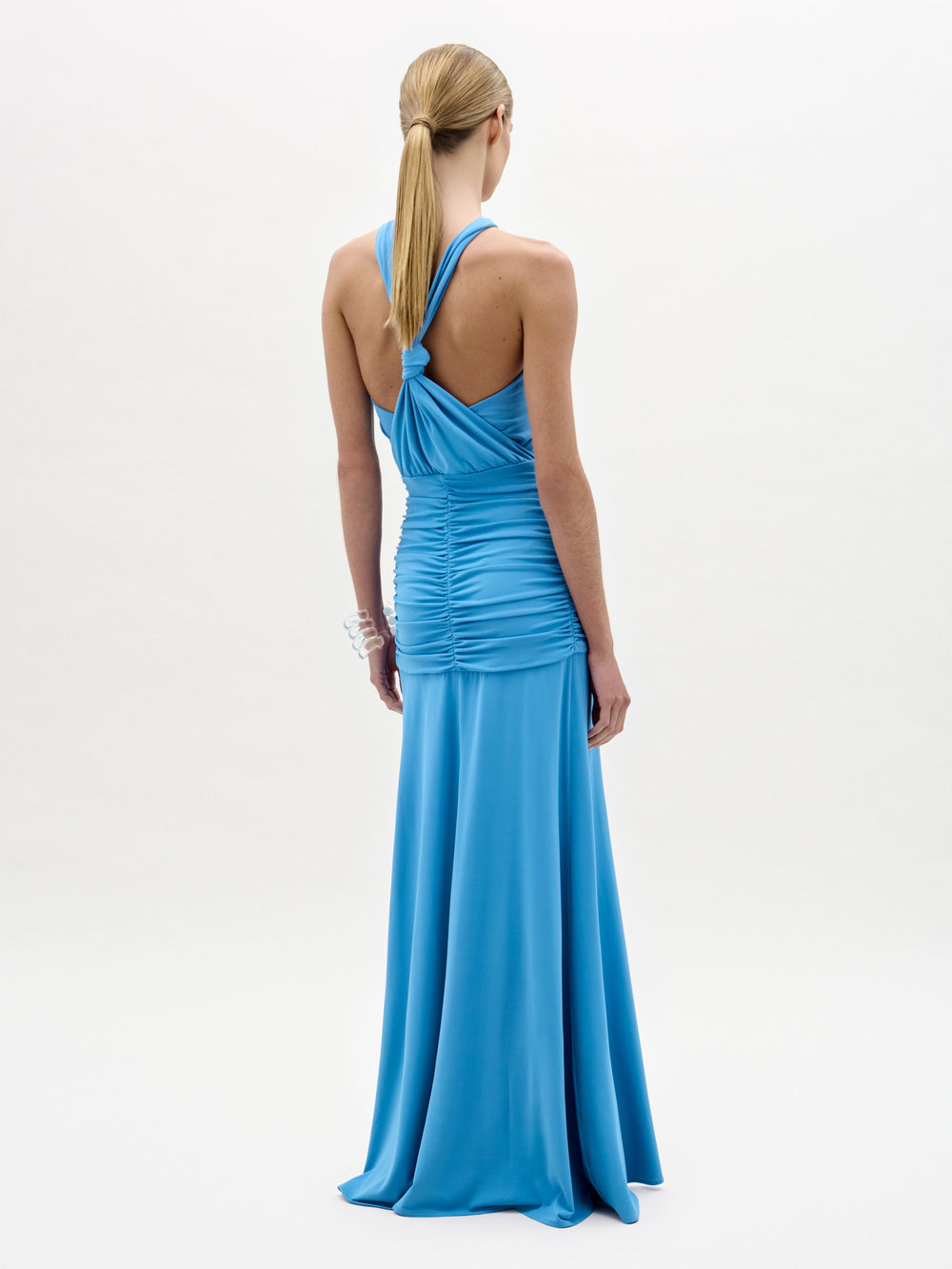 A person with straight blonde hair in a low ponytail is seen from behind, wearing the Jedda Dress by SVT DESIGNS—a sleeveless, floor-length blue maxi dress with ruched detailing, open back, and fitted silhouette against a plain white background.