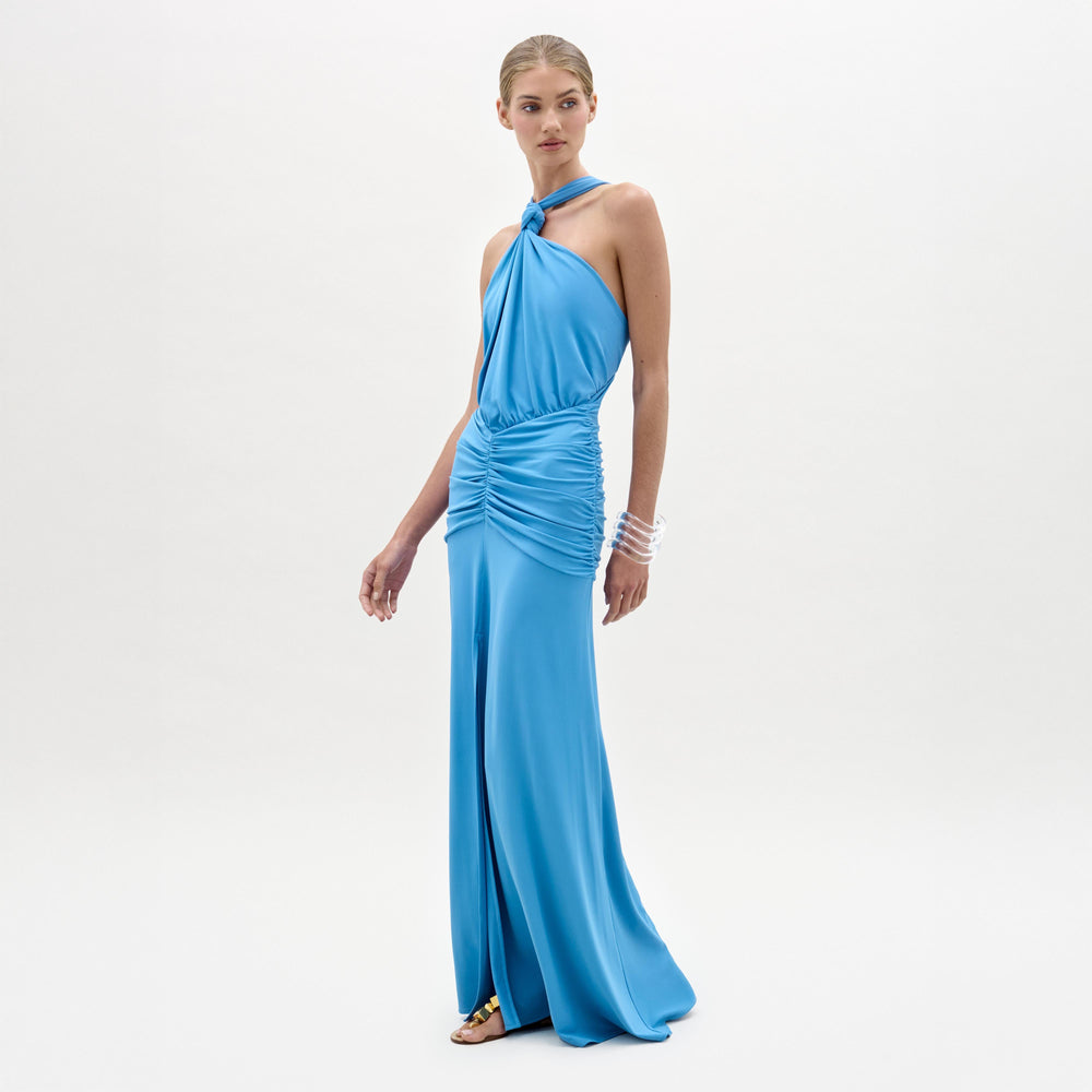 A woman stands against a white background wearing the sleeveless, halter-neck blue Jedda Dress by SVT DESIGNS, featuring gathered fabric at the waist and hips for a fitted silhouette. She has her hair tied back and wears flat sandals and bracelets.