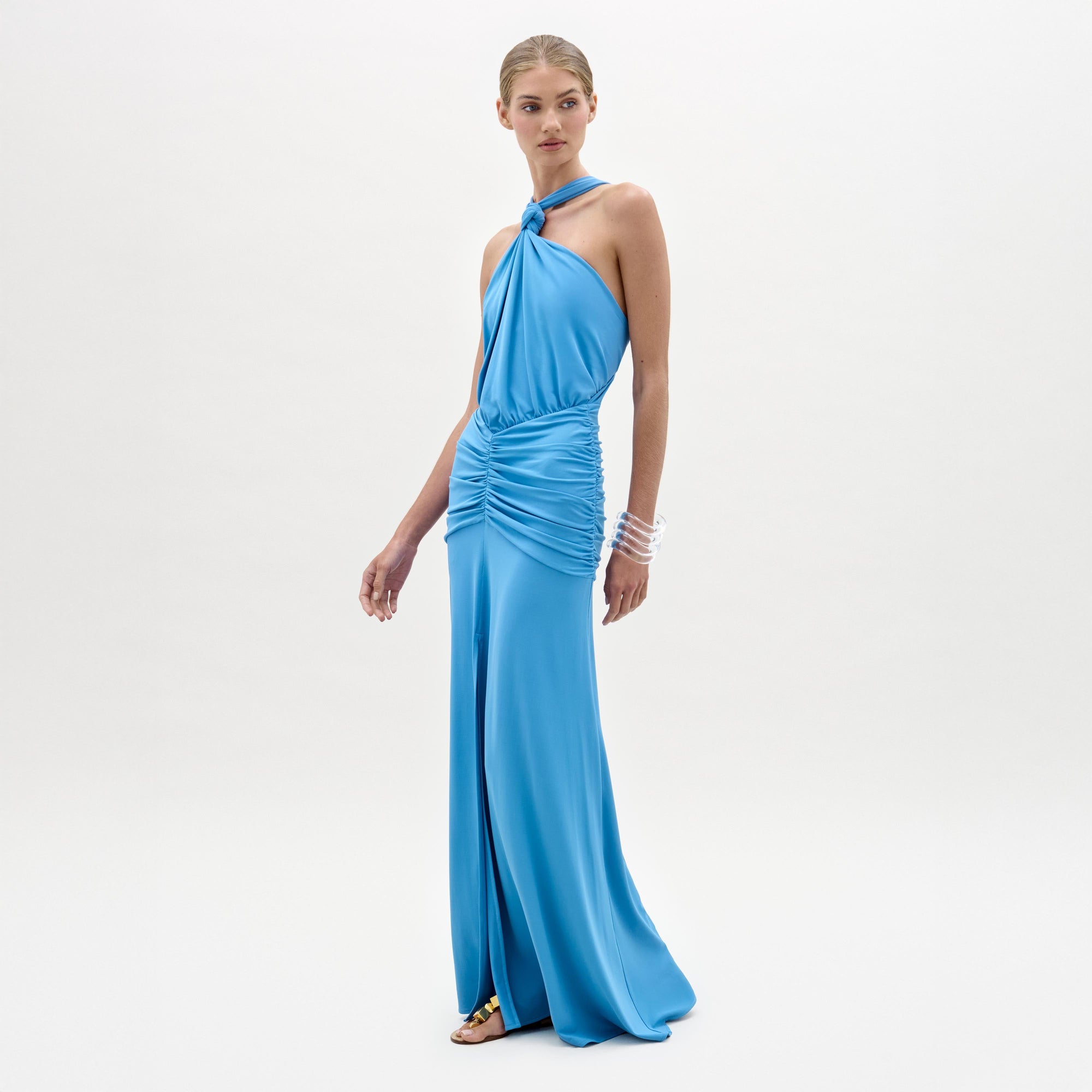 A woman stands against a white background wearing the sleeveless, halter-neck blue Jedda Dress by SVT DESIGNS, featuring gathered fabric at the waist and hips for a fitted silhouette. She has her hair tied back and wears flat sandals and bracelets.