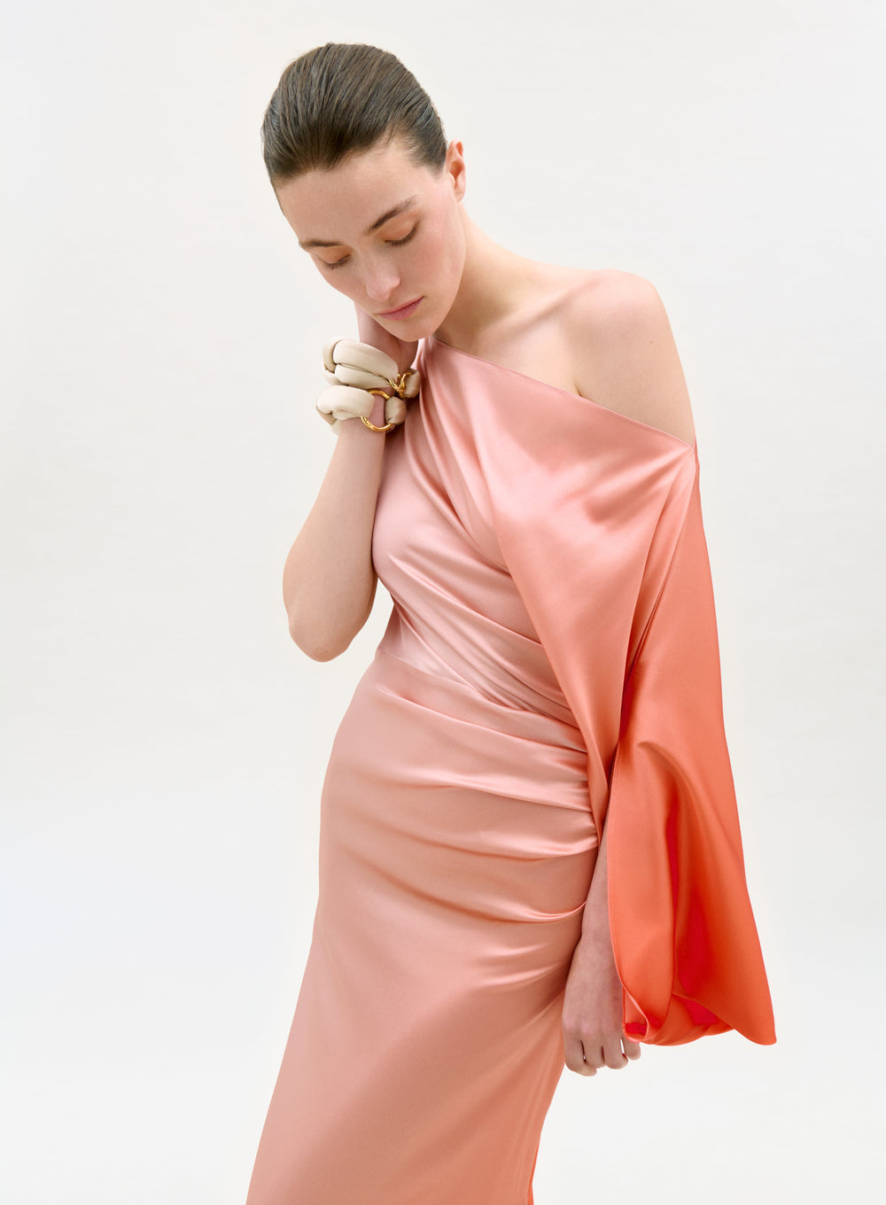 A woman models the Frankee Dress—a shiny, peach satin one-shoulder gown—against a plain background, looking down thoughtfully with her hair pulled back and a soft, elegant expression.