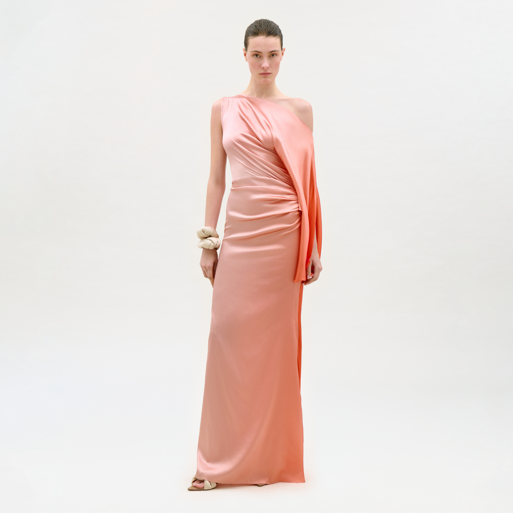 A woman stands against a white background wearing the Frankee Dress, a long, peach one-shoulder satin gown with draped detailing. She accessorizes with a bracelet, has her hair pulled back, and looks directly at the camera.