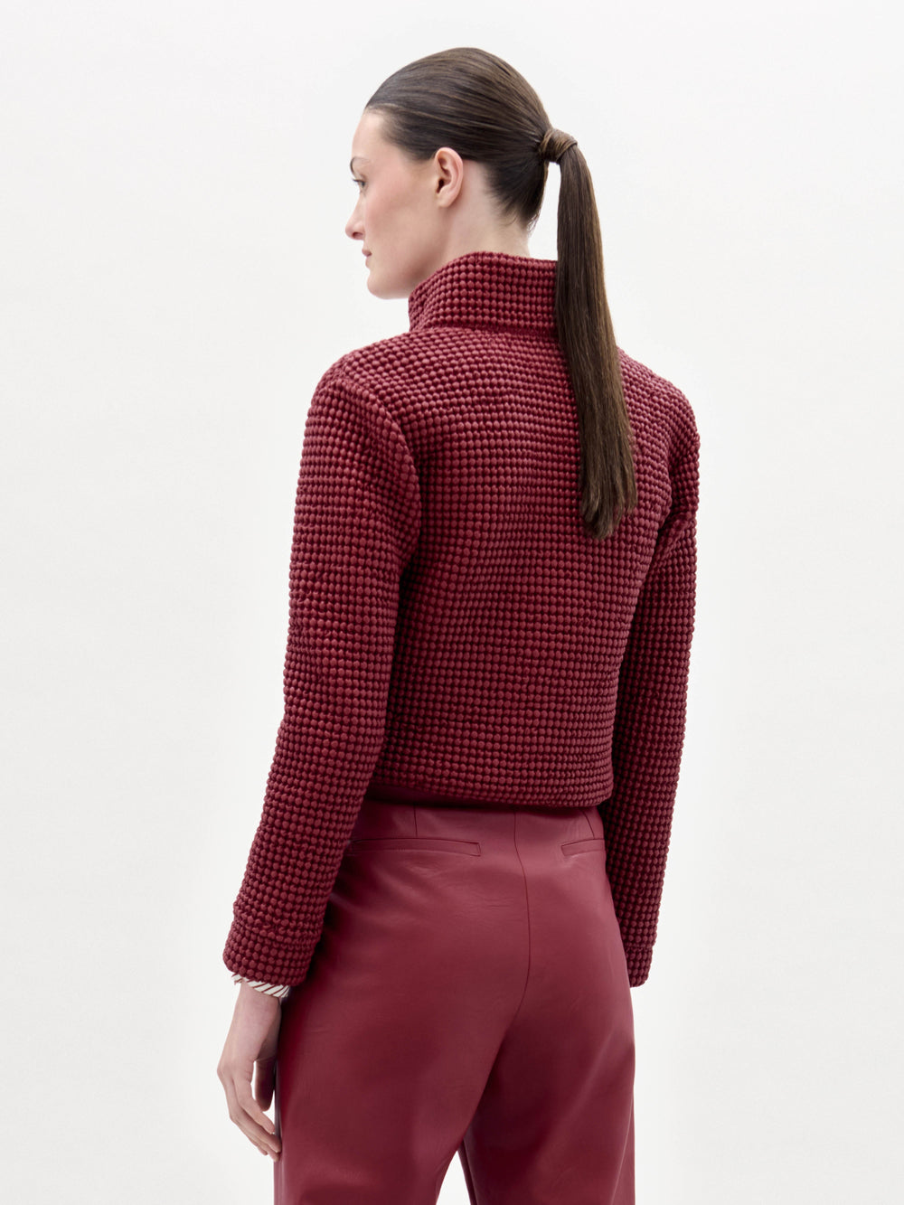 A woman stands against a plain background wearing the Zalta Jacket, a textured red jacket paired with a striped shirt, red pants, and a large black belt featuring an ornate buckle.