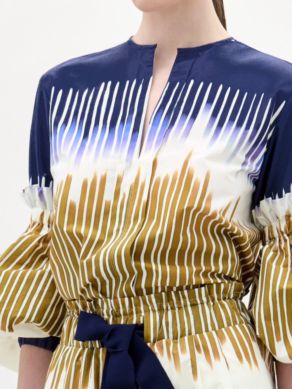A woman wears the Lucaya Blouse by SVT DESIGNS, made from 100% cotton with navy, white, and brown vertical stripes, a round neckline with a small front slit, voluminous sleeves, and a navy waist belt. Her hair is pulled back.
