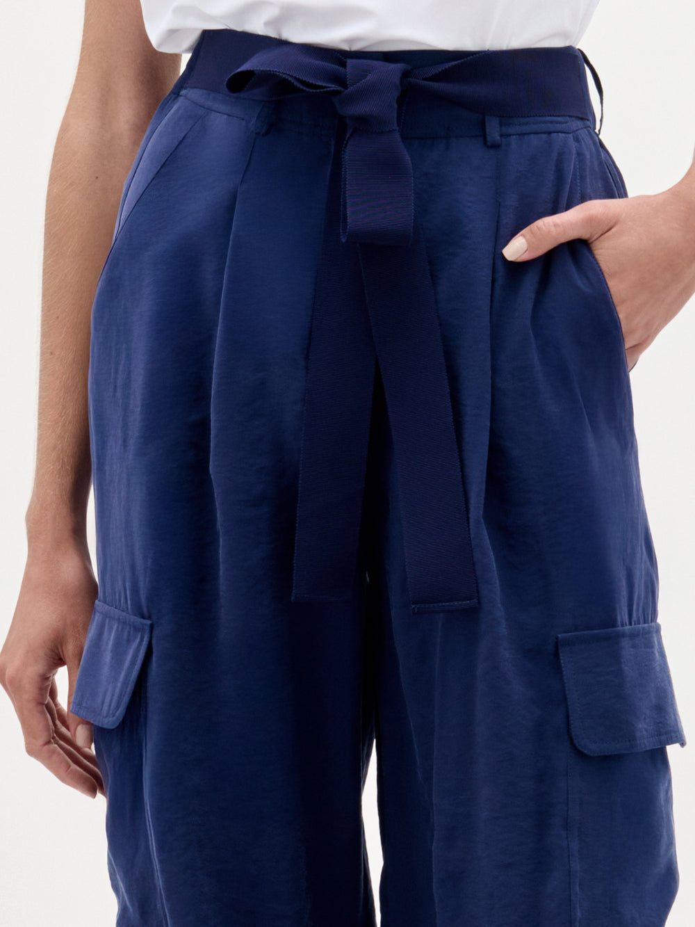 A person wears SVT DESIGNS' Darnell Pant—high waist navy blue satin cargo pants with side pockets and matching belt—with a white top. Right hand in pocket; only torso and upper legs are visible.