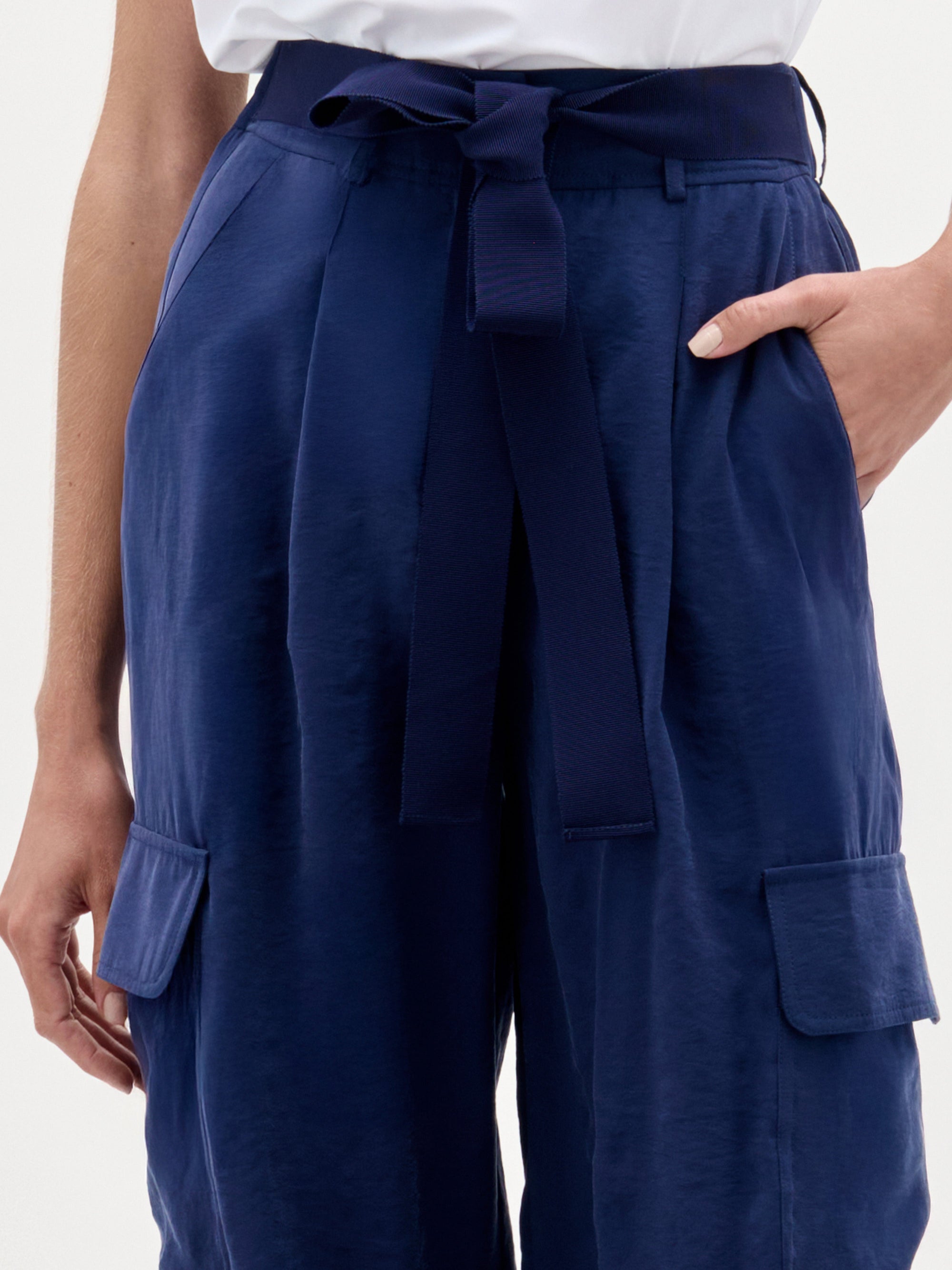 A person wears SVT DESIGNS' Darnell Pant—high waist navy blue satin cargo pants with side pockets and matching belt—with a white top. Right hand in pocket; only torso and upper legs are visible.