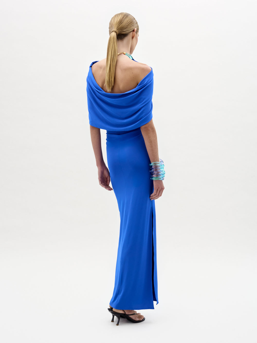 A woman with a low ponytail and black heels wears the SVT DESIGNS Odele Dress—a floor-length blue maxi with a halter neckline and side slit—paired with turquoise bangles, against a plain white background.