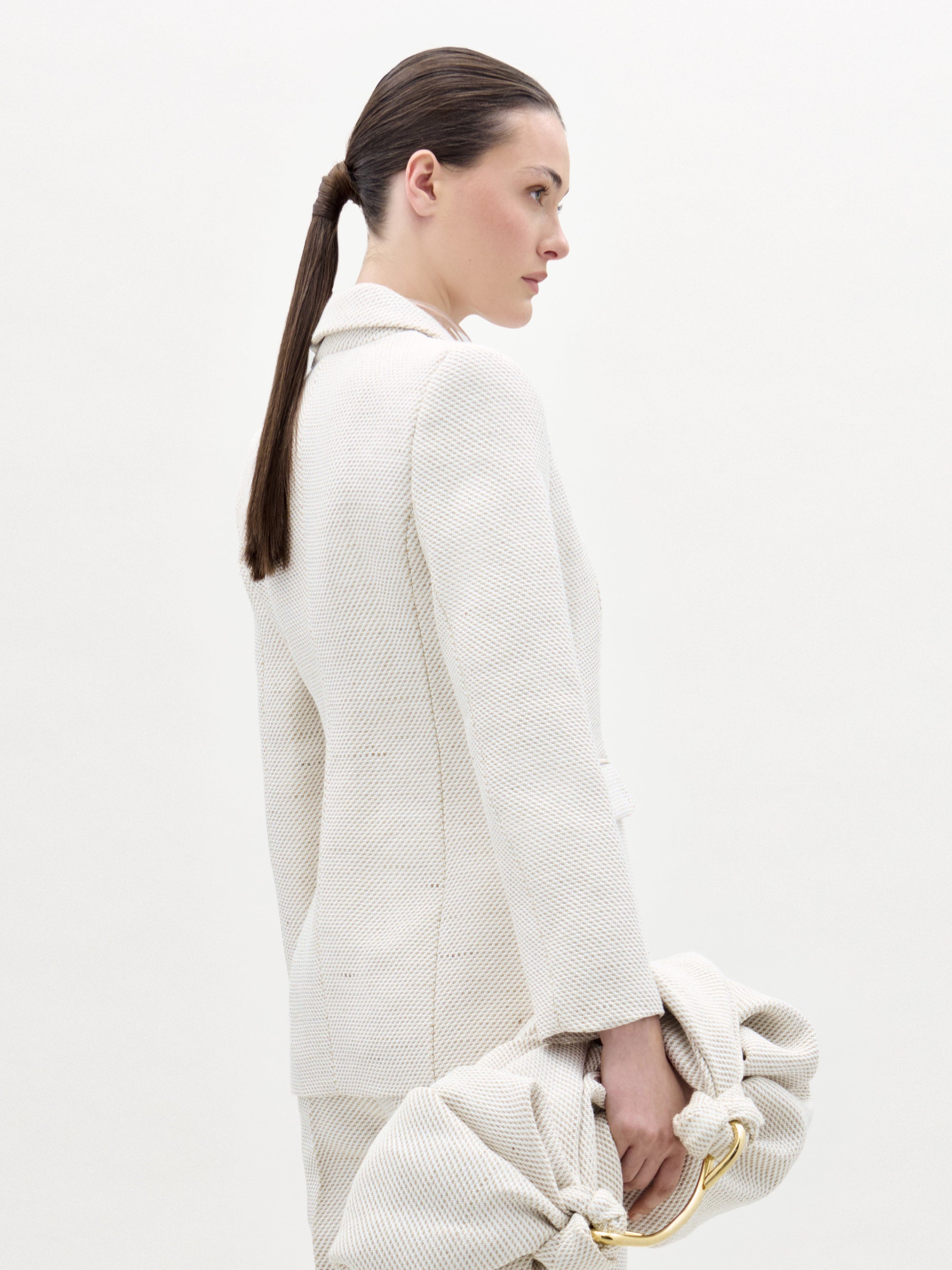 A woman with dark hair in a low ponytail stands sideways wearing the SVT DESIGNS Rebeca Jacket, a textured light blazer, and holds a matching large gathered clutch against a plain light background.