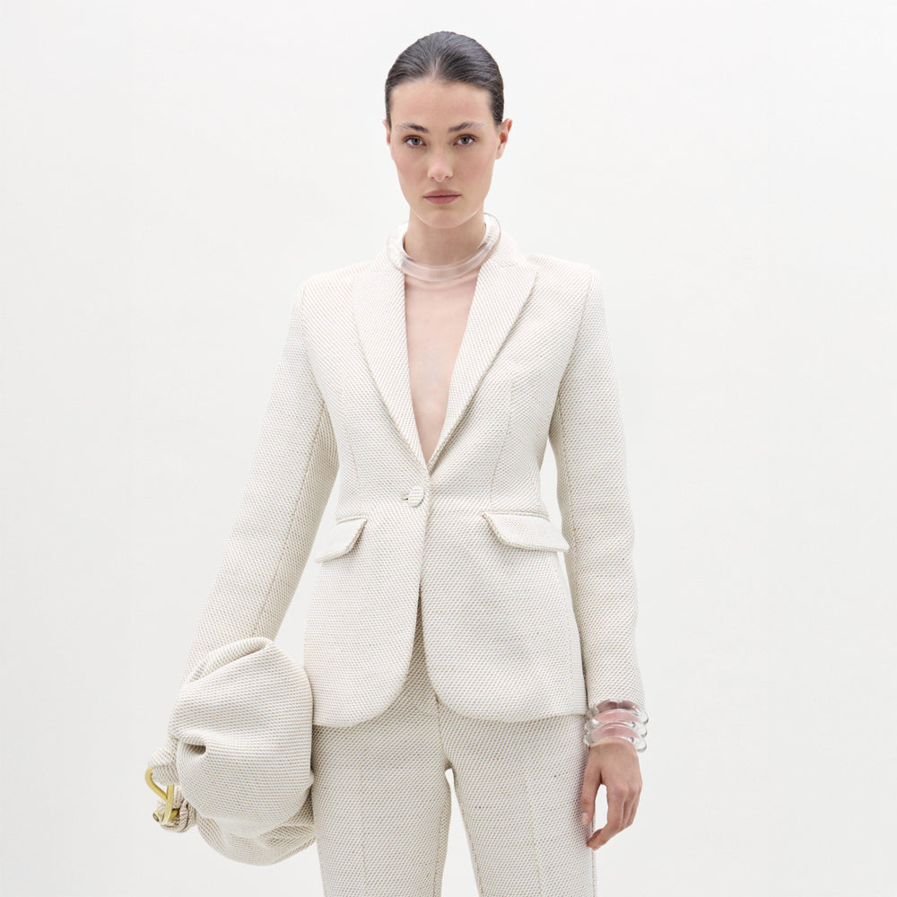 Against a plain white background, a person wears SVT DESIGNS' textured off-white Rebeca Jacket with matching pants, accessorizing with translucent bracelets and a large, gathered bag in their left hand.