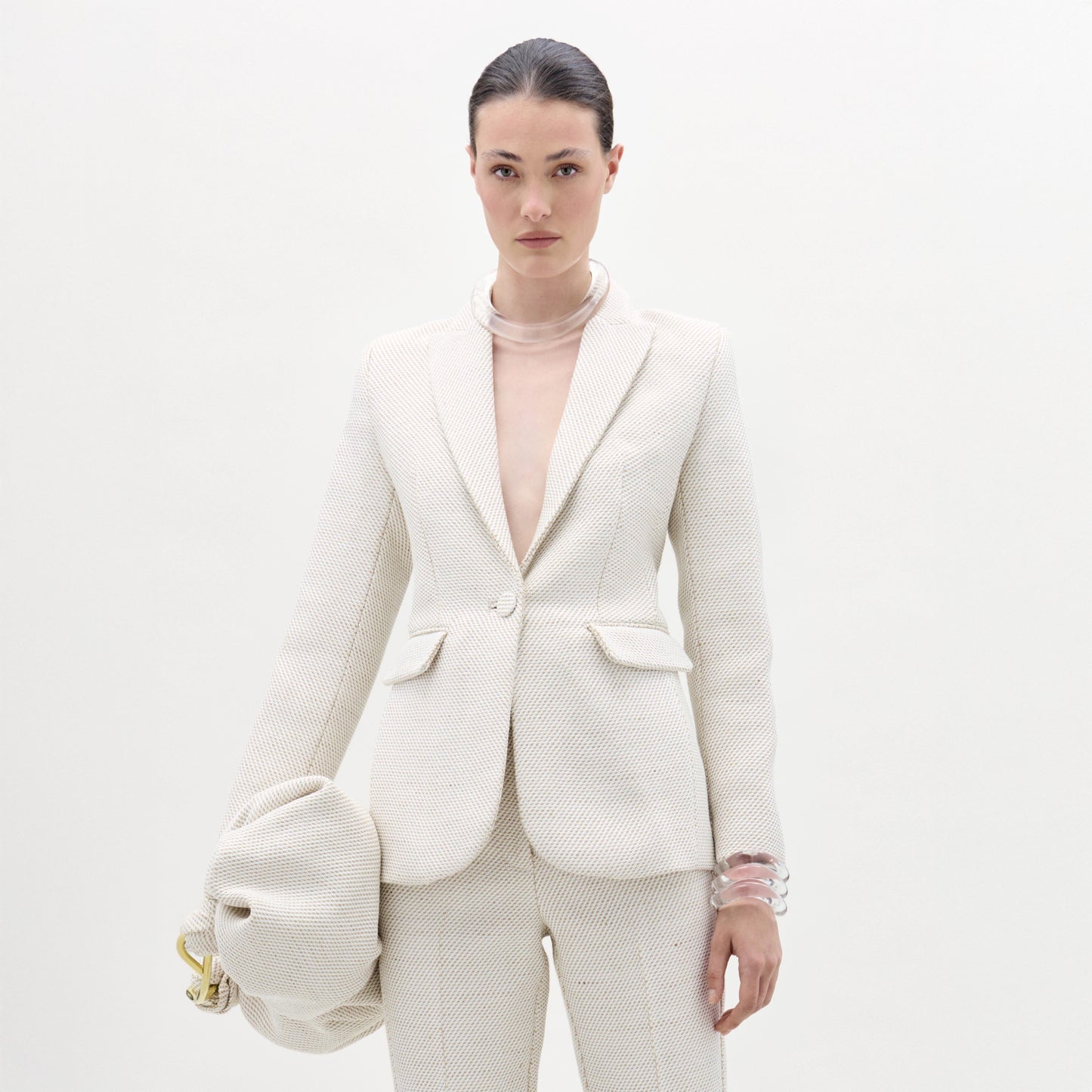 Against a plain white background, a person wears SVT DESIGNS' textured off-white Rebeca Jacket with matching pants, accessorizing with translucent bracelets and a large, gathered bag in their left hand.