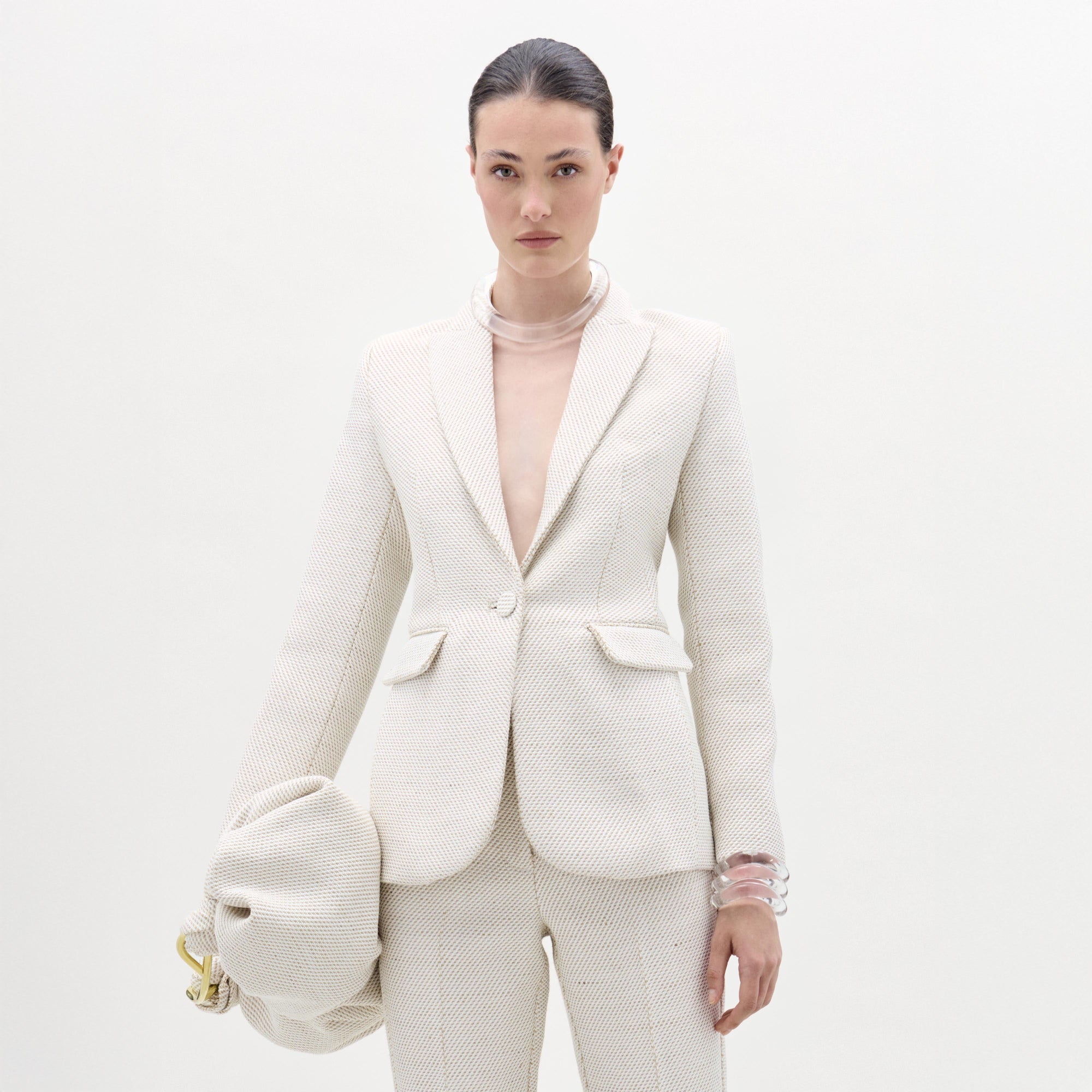 Against a plain white background, a person wears SVT DESIGNS' textured off-white Rebeca Jacket with matching pants, accessorizing with translucent bracelets and a large, gathered bag in their left hand.