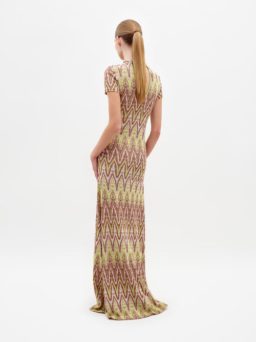 A person standing and posing in the Yara Dress Zigzag Neon, a floor-length dress with a vibrant zigzag design, adorned with large bracelet accessories and wearing flat sandals, against a white background. Pre-order now to ensure your Yara Dress Zigzag Neon ships by February 15th.