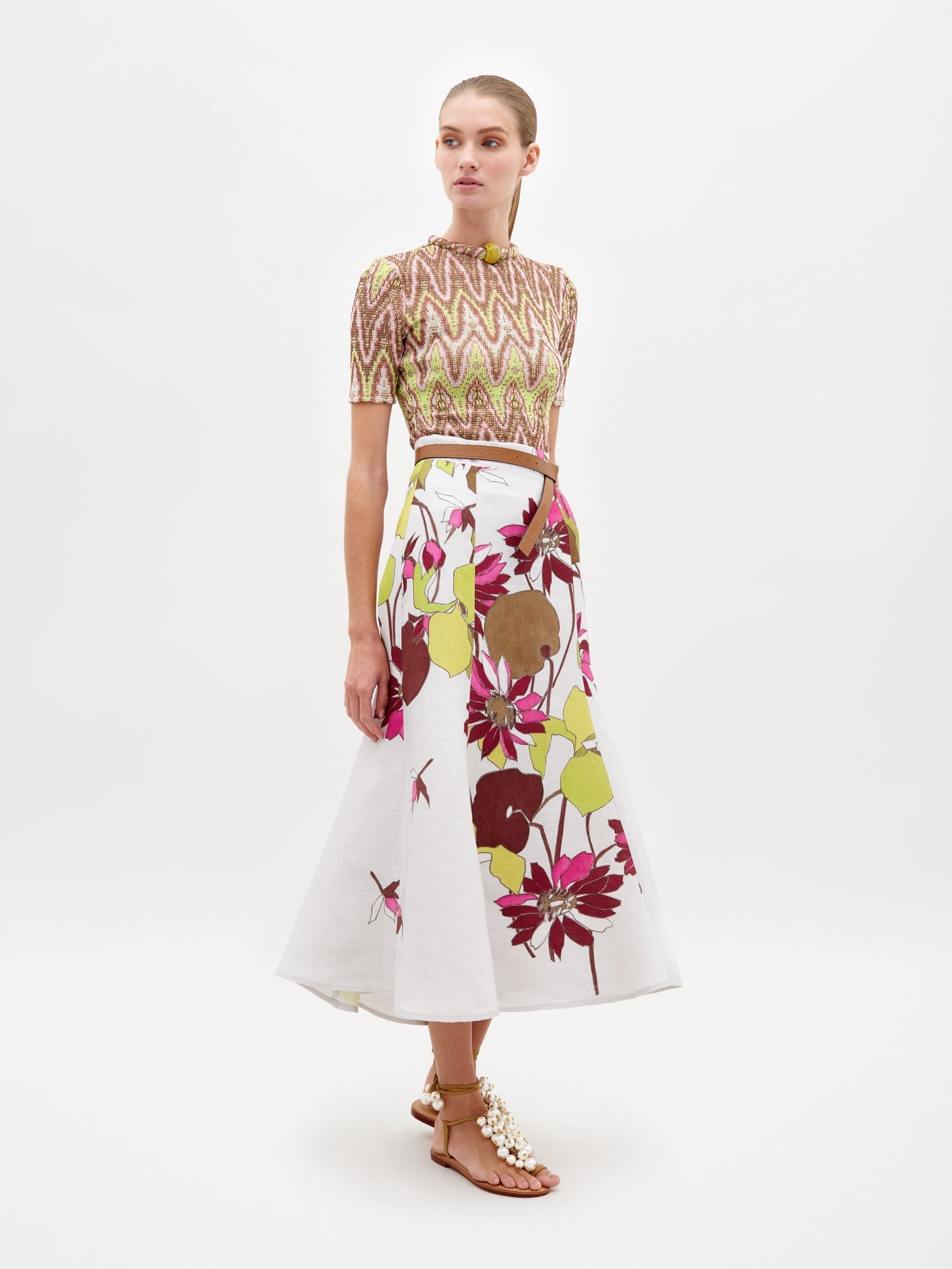 A person standing in a patterned top and the Erin Skirt Botanical Multifloral paired with sandal heels against a plain white background, ready for pre-order. Expected ship by date is February 15th, 2025.