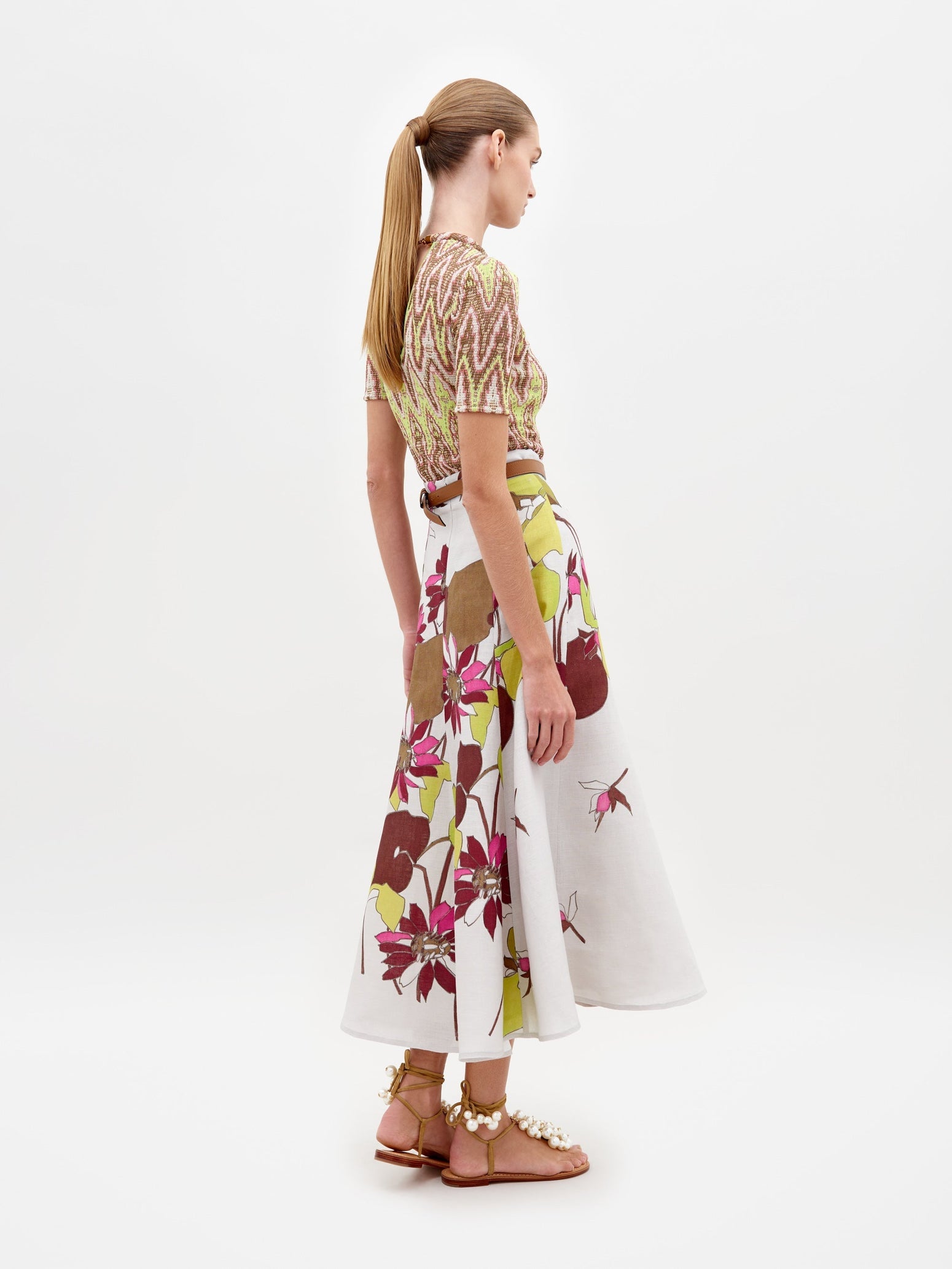 A person standing in a patterned top and the Erin Skirt Botanical Multifloral paired with sandal heels against a plain white background, ready for pre-order. Expected ship by date is February 15th, 2025.