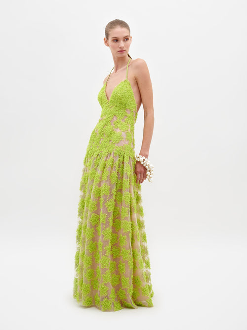 A woman stands wearing the Danae Dress Lime, a stunning floor-length, lime green halter dress featuring a textured floral pattern. She accessorizes with a bracelet on her wrist and poses against a plain white background, exuding elegance for 2025. Ensure delivery by the ship-by date of February 15th.