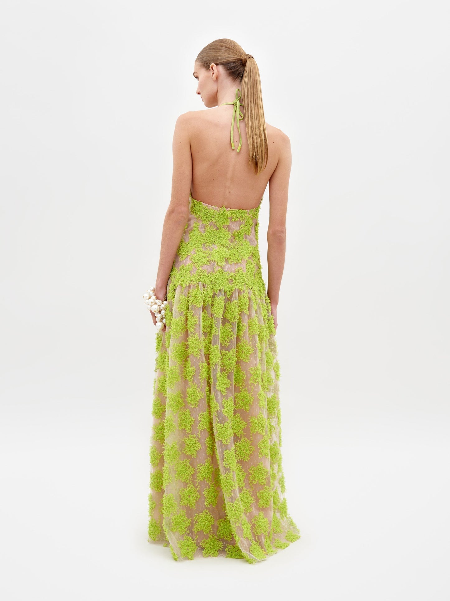 A woman stands wearing the Danae Dress Lime, a stunning floor-length, lime green halter dress featuring a textured floral pattern. She accessorizes with a bracelet on her wrist and poses against a plain white background, exuding elegance for 2025. Ensure delivery by the ship-by date of February 15th.