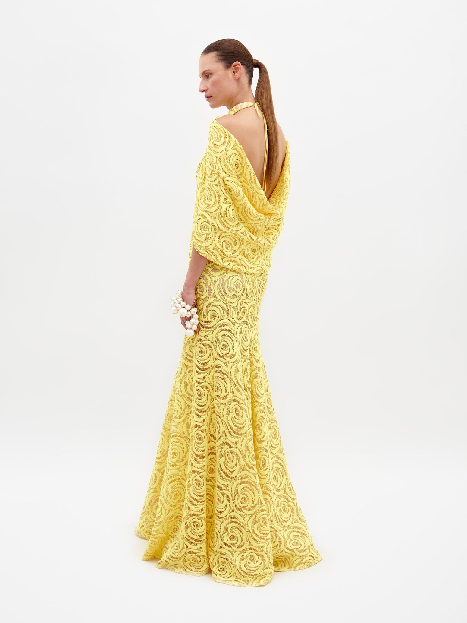 Person wearing a Salvina Dress Tuscany Yellow, adorned with a high neckline and cape-like sleeves, holding a pearl bracelet on their wrist, eagerly awaiting the pre-order ship date for 2025.