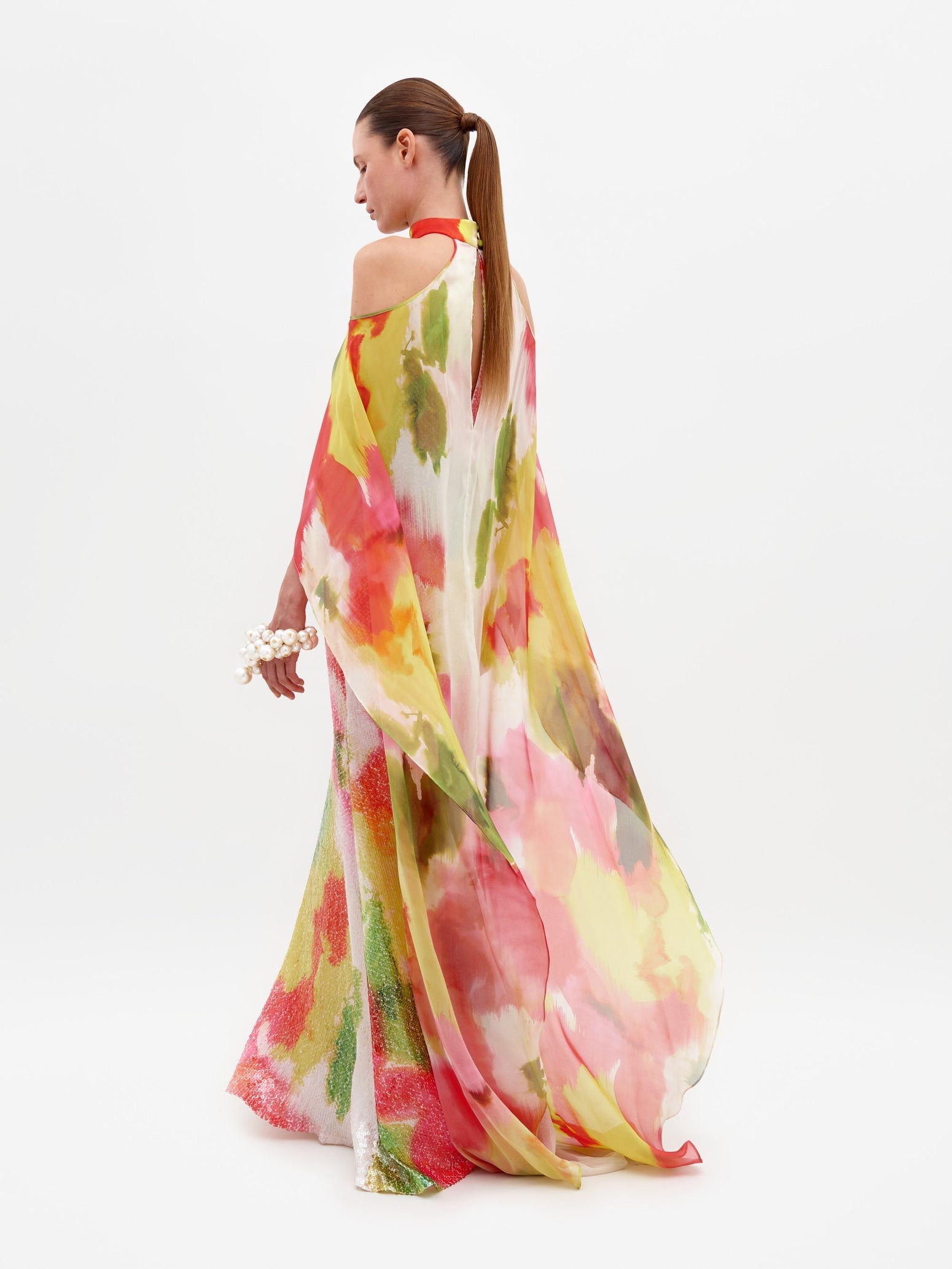 A person is standing in the Serenela Dress Spring Blooms, a colorful, flowing dress with a halter neck and floral patterns, holding a small white flower bouquet. The dress is ready to be pre-ordered for the February 15th ship-by date.