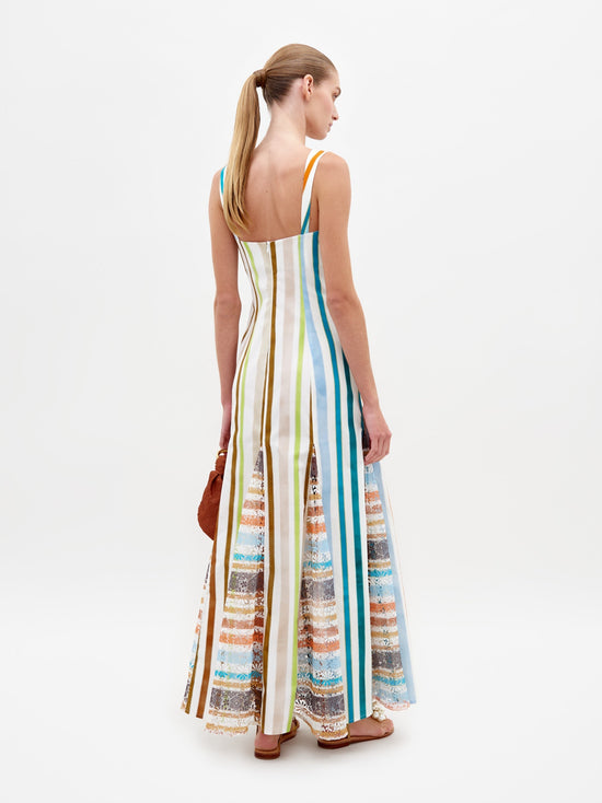 A woman stands wearing the Ofra Dress Multi Color Stripes, a sleeveless outfit featuring vibrant stripes and floral details, while holding an orange bag. The background is plain white. Please note this item is available for pre-order and will ship by February 15th, 2025.