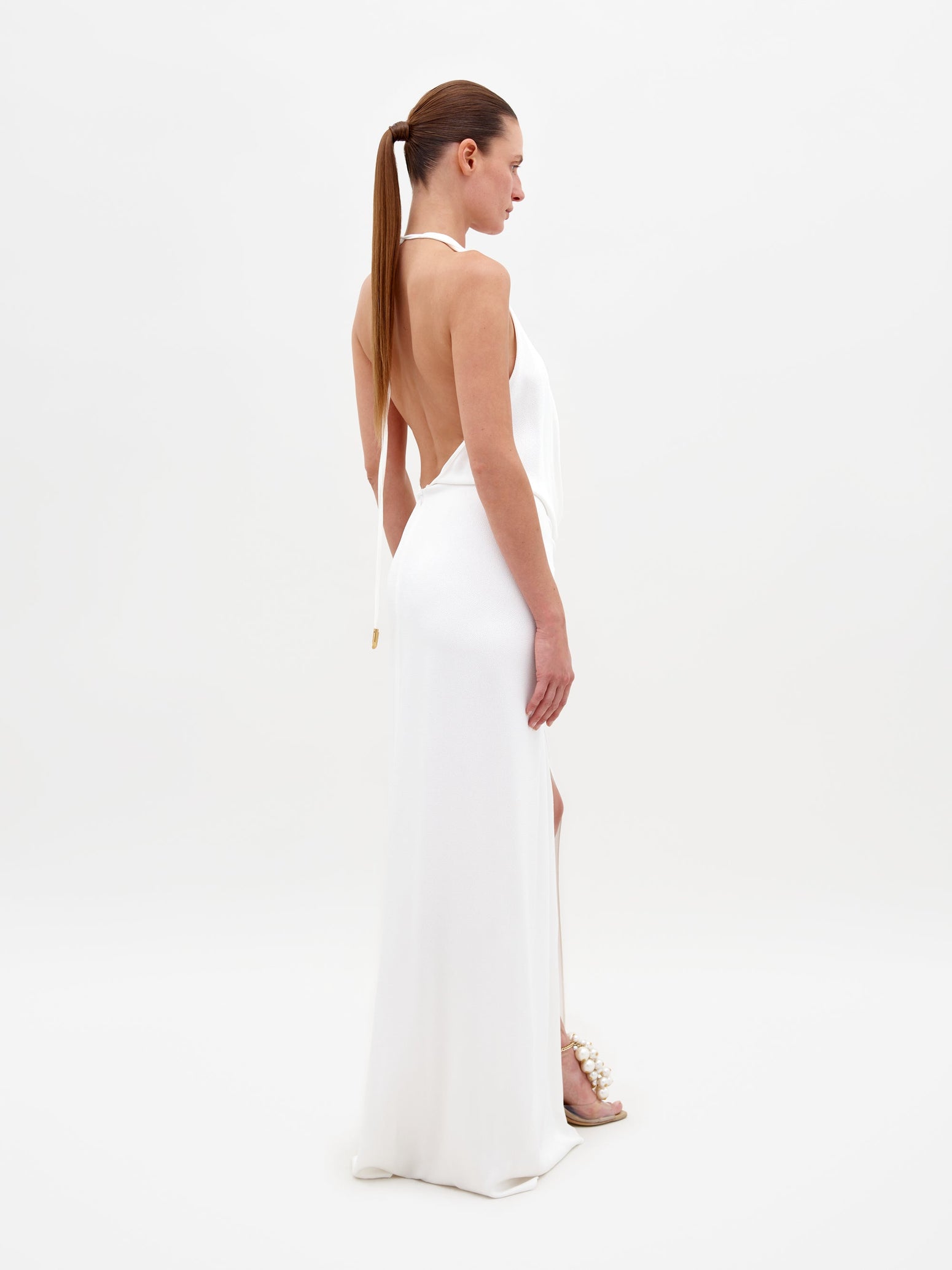 Person standing against a white background, wearing the Torgiano Dress White, which features a sleeveless halter design with a knot detail at the waist and a front slit. Pre-order now to ensure your dress is shipped by February 15th, 2025.