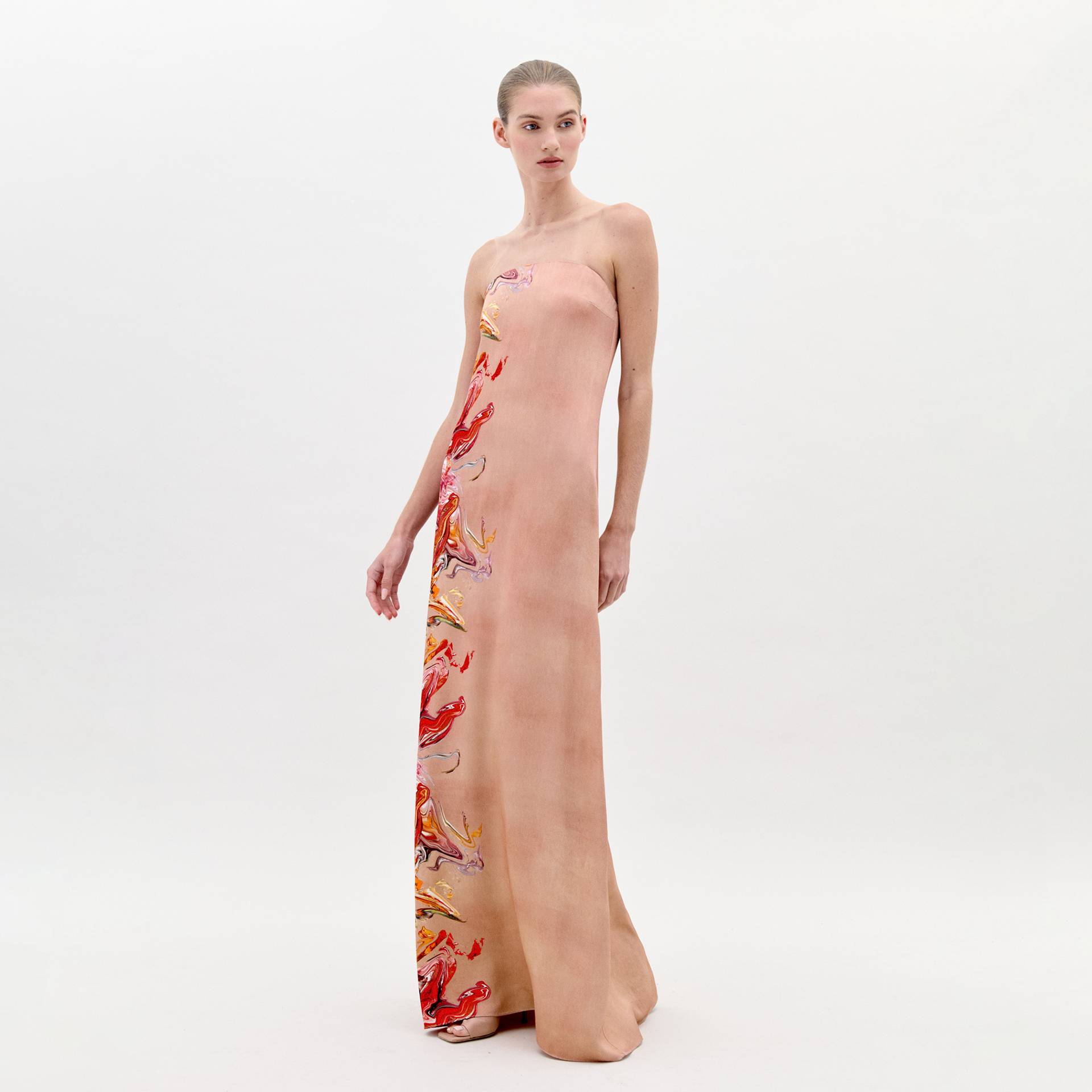 A woman stands in a long, strapless Adalgisa Dress Abstract Coral with red, orange, and yellow floral accents on the side against a plain white background. This stunning piece is available for pre-order now with an estimated ship-by date of February 1st, 2025.