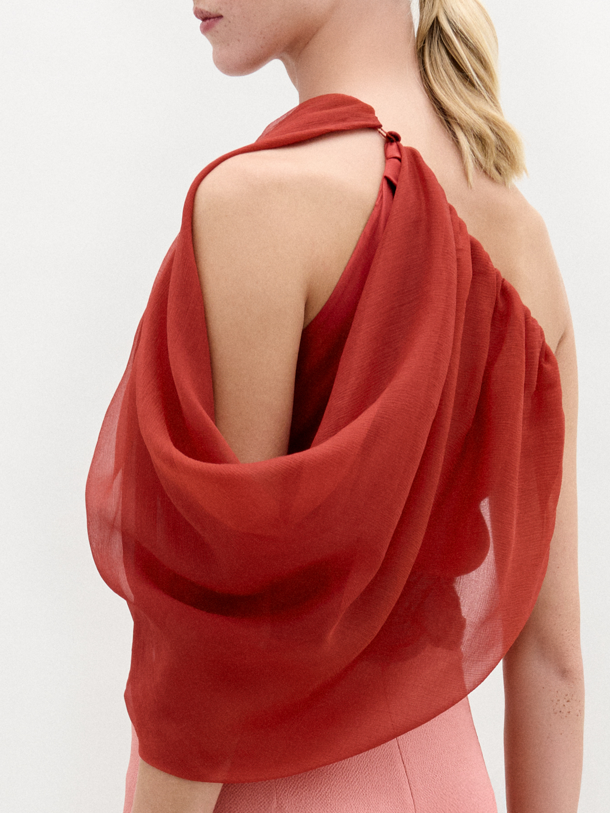 A woman wears the Adil Blouse in red with a one-shoulder neckline, paired with pink high-waisted pants, a wide belt with a large gold buckle, and gold bracelets, standing against a plain white background.