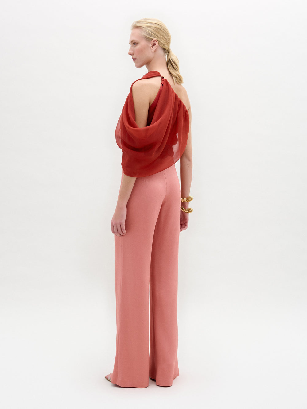 A woman stands in high-waisted pink Palermo Pants, crafted from luxurious Italian fabric, paired with a red one-shoulder top, gold belt, gold bracelets, and open-toe shoes against a white background.
