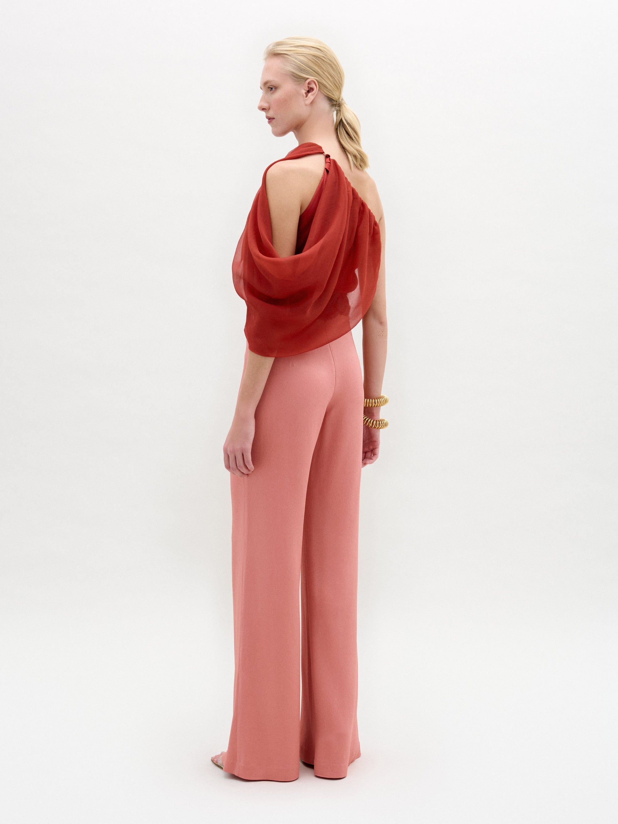 A woman stands in high-waisted pink Palermo Pants, crafted from luxurious Italian fabric, paired with a red one-shoulder top, gold belt, gold bracelets, and open-toe shoes against a white background.