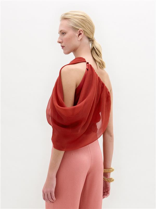 A person with blond hair in a low ponytail models the SVT DESIGNS Adil Blouse—a sheer, red, draped one-shoulder sleeveless top—paired with high-waisted pink pants and two gold bangles, standing against a plain white background.