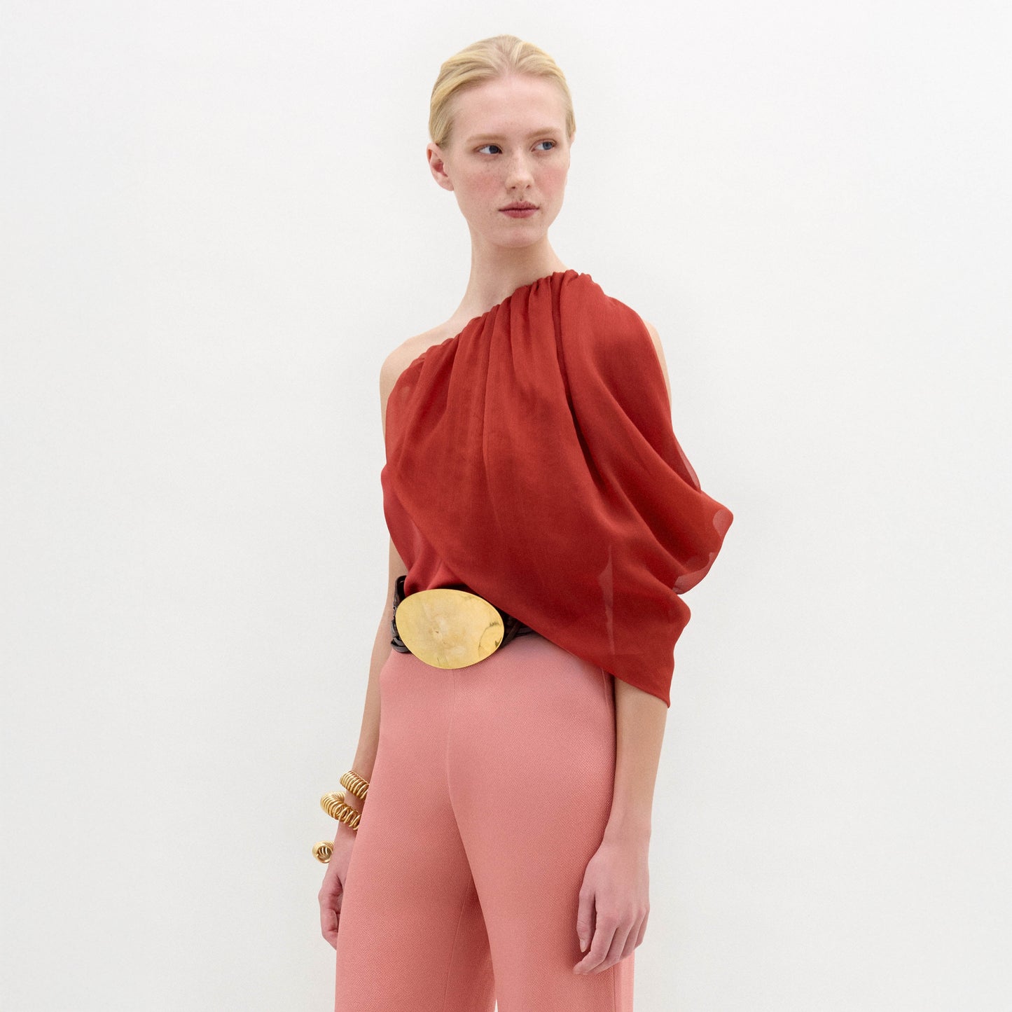 A woman wears the Adil Blouse in red with a one-shoulder neckline, paired with pink high-waisted pants, a wide belt with a large gold buckle, and gold bracelets, standing against a plain white background.